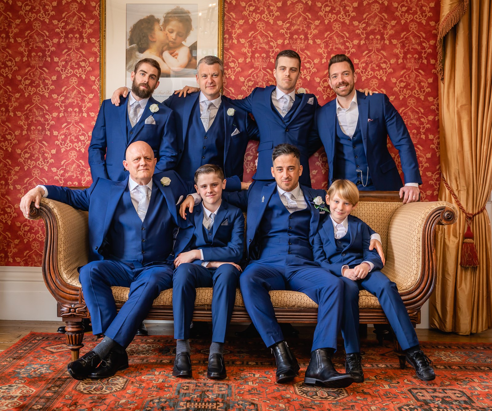 The groom and his groomsmen posed on a sofa against red damask wallpaper at Fennes