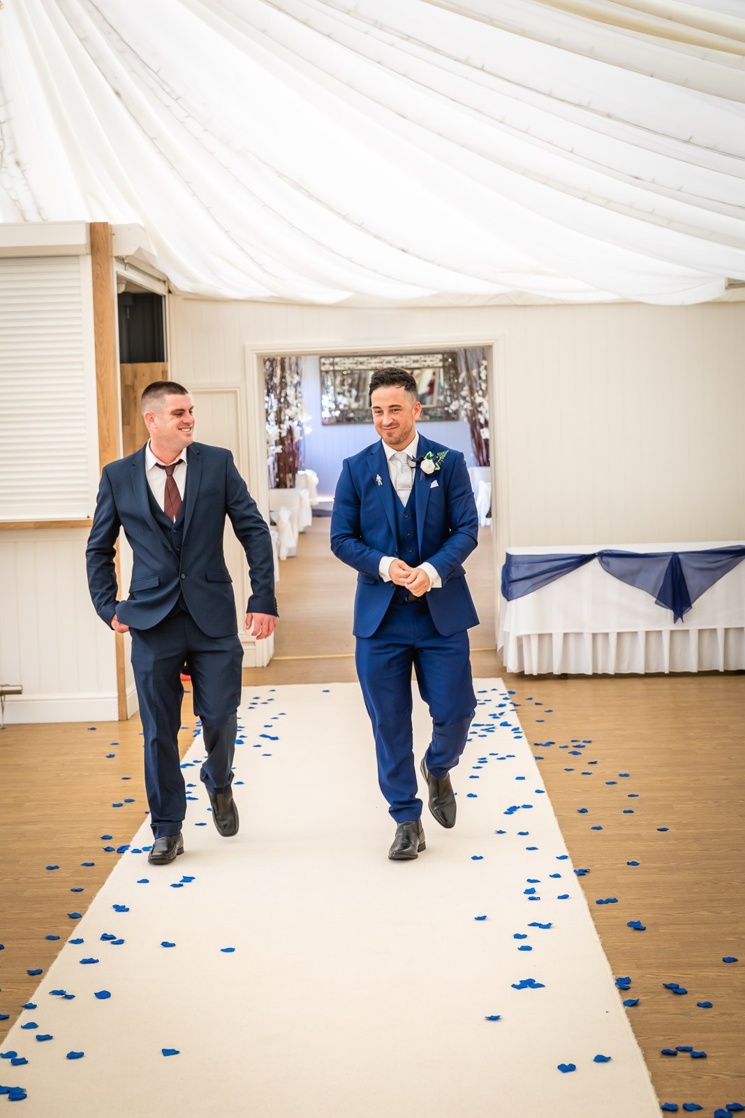 Groom and a guest walking down the petal-strewn aisle in the draped ceremony room