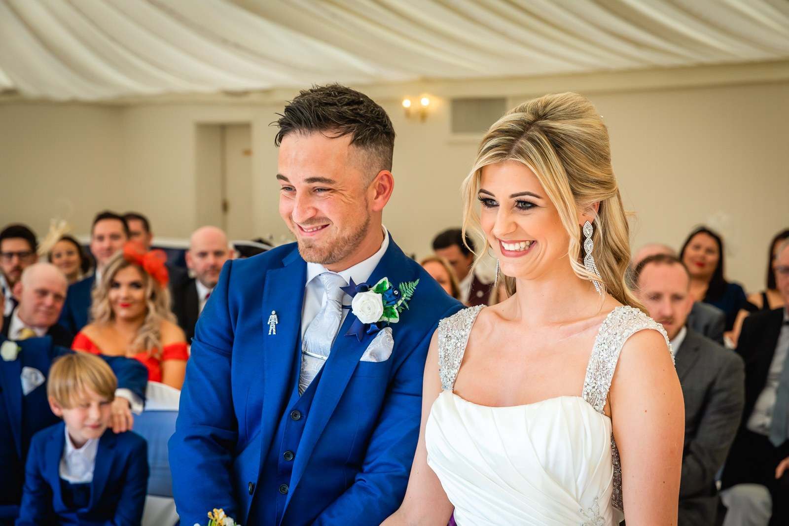 Bride and groom smiling side by side during the ceremony as guests watch