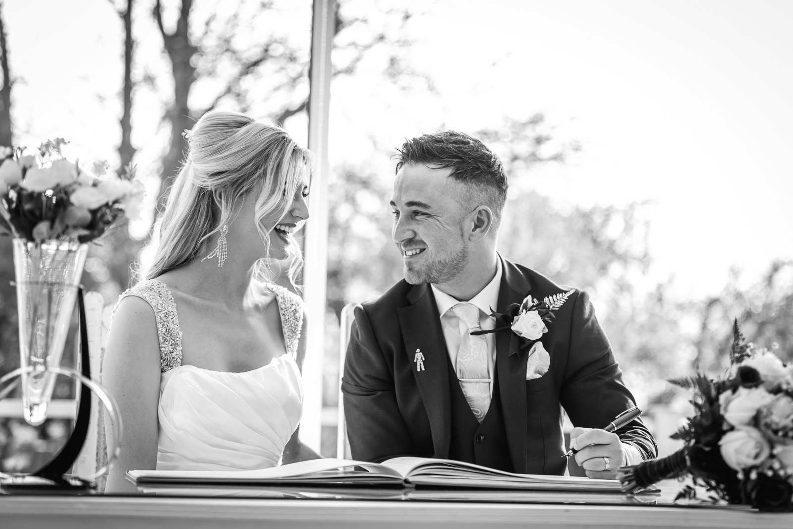 Black-and-white of the couple smiling at each other while signing the register
