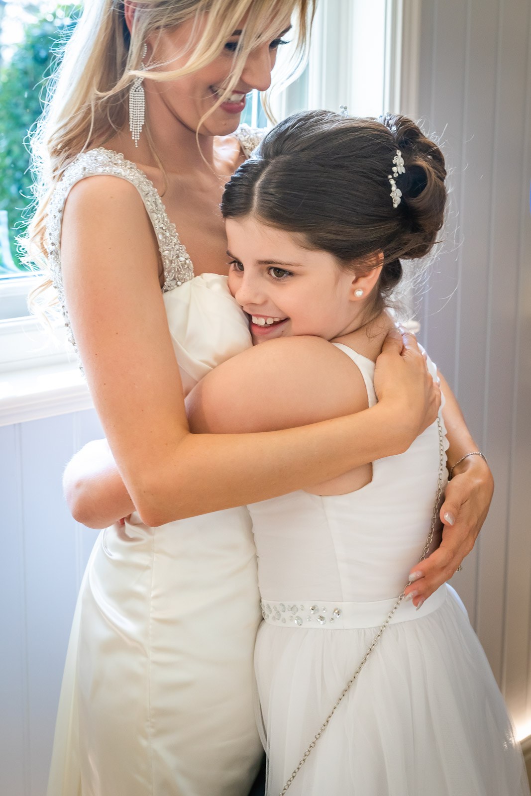 The bride hugging a young flower girl by a sunlit window