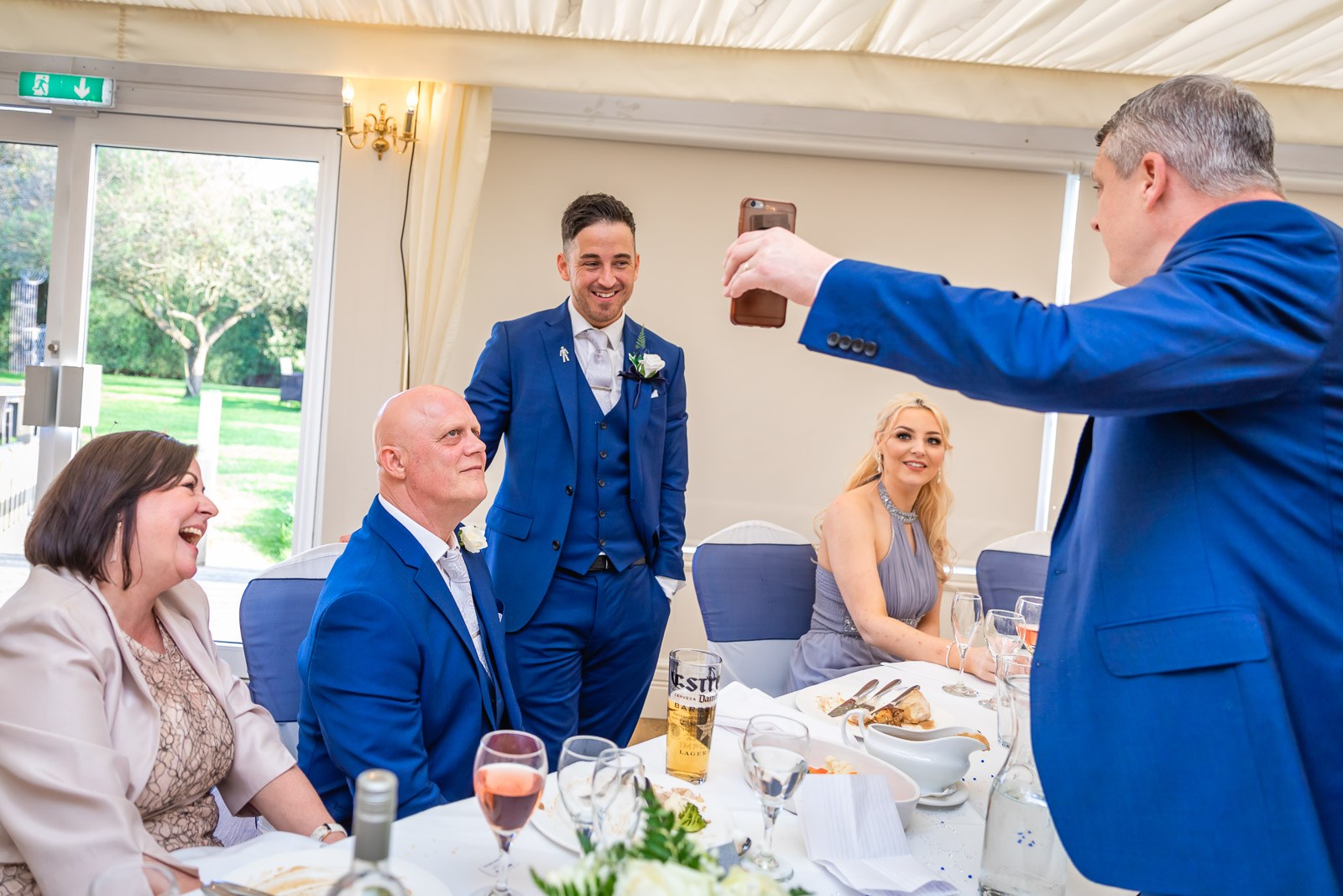 A guest holding up a phone to photograph the groom as others laugh