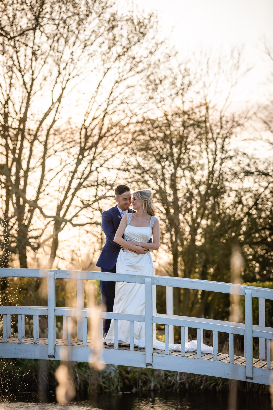 The couple embracing on the white footbridge at Fennes in golden evening light