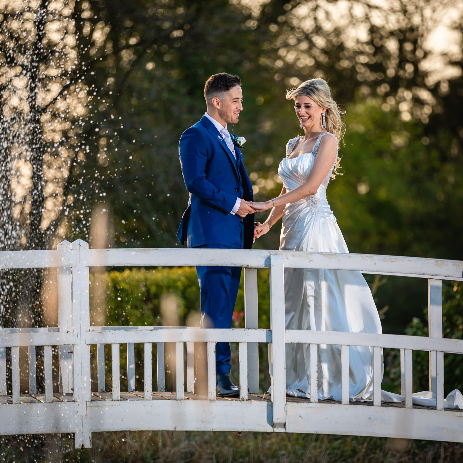 The couple holding hands on the footbridge at Fennes, the lake fountain spraying behind