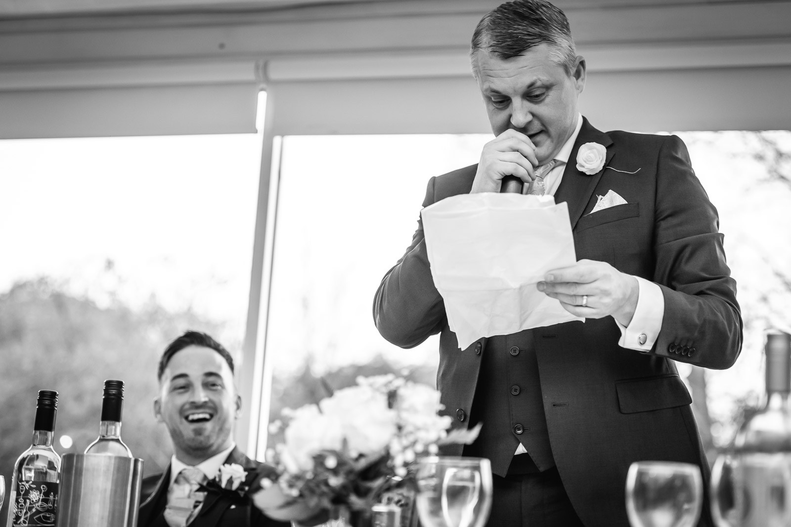 Black-and-white of a groomsman reading his speech as the groom laughs