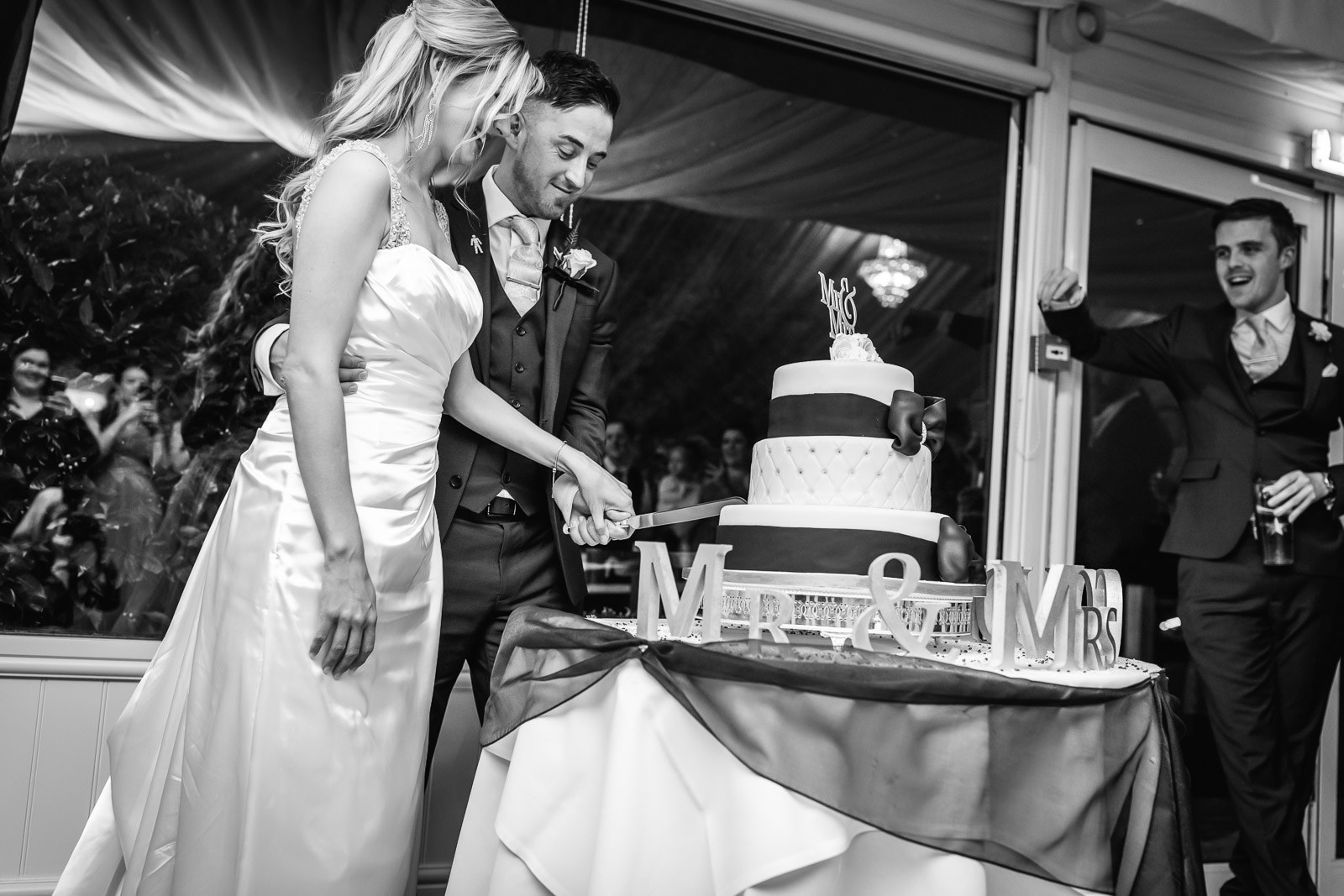 Black-and-white of the couple cutting their cake as guests watch