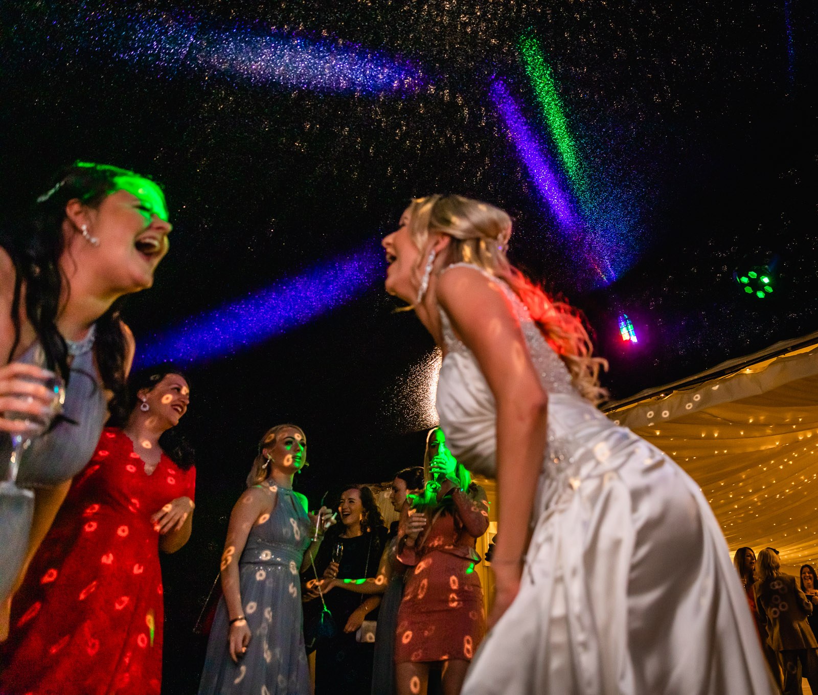 The bride dancing with a bridesmaid, drinks in hand, under the lights