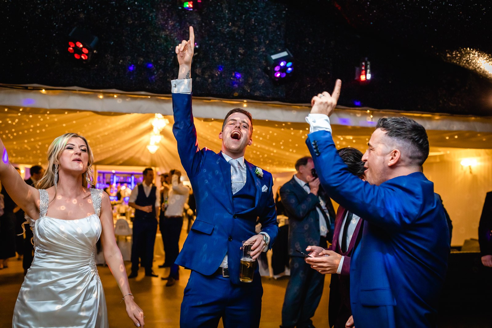 Bride and groom dancing, the groom singing with an arm raised, in the fairy-lit marquee