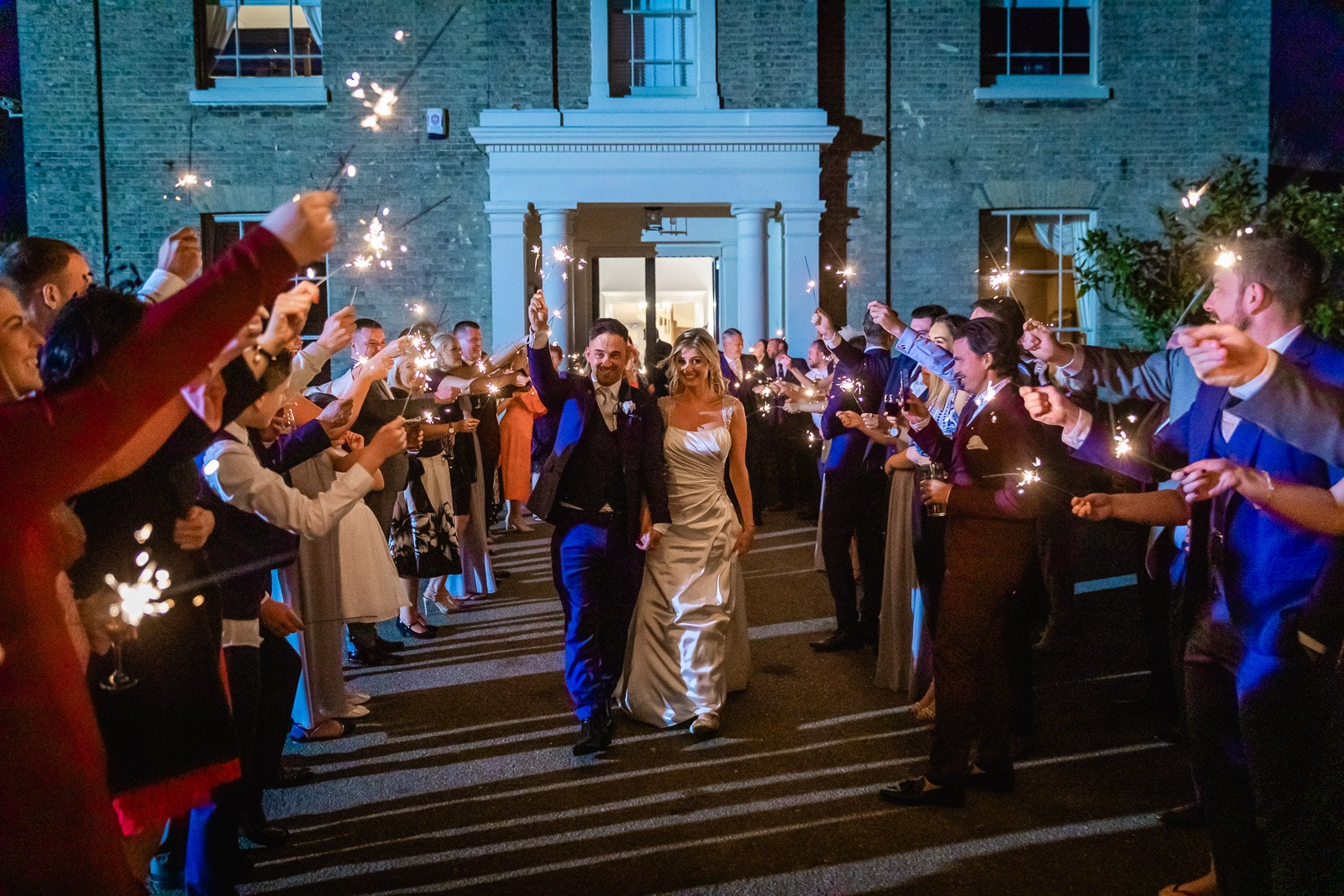 Newlyweds walking a sparkler tunnel of guests outside the lit house at Fennes