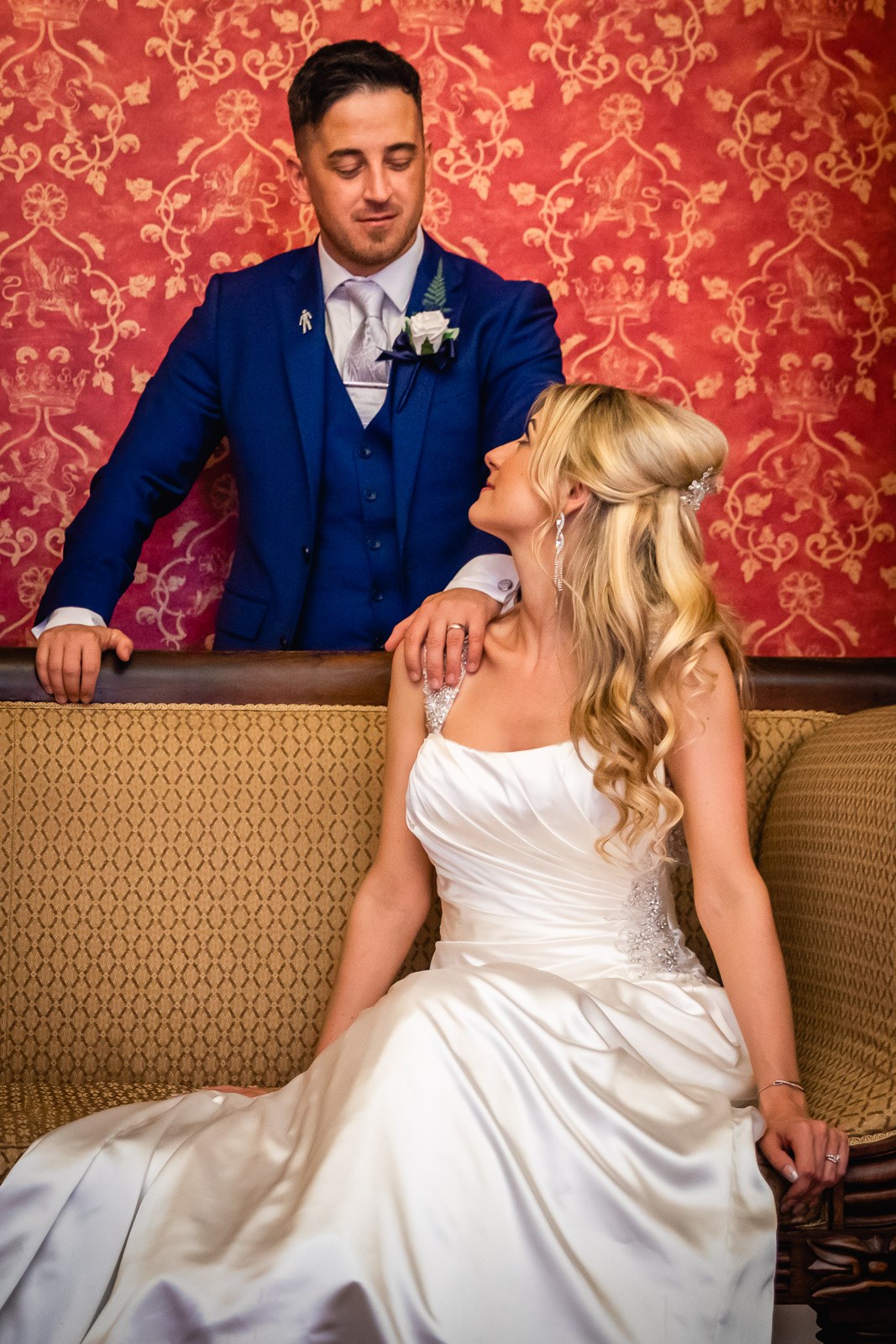 The groom leaning over the sofa toward the bride in the red damask lounge at Fennes