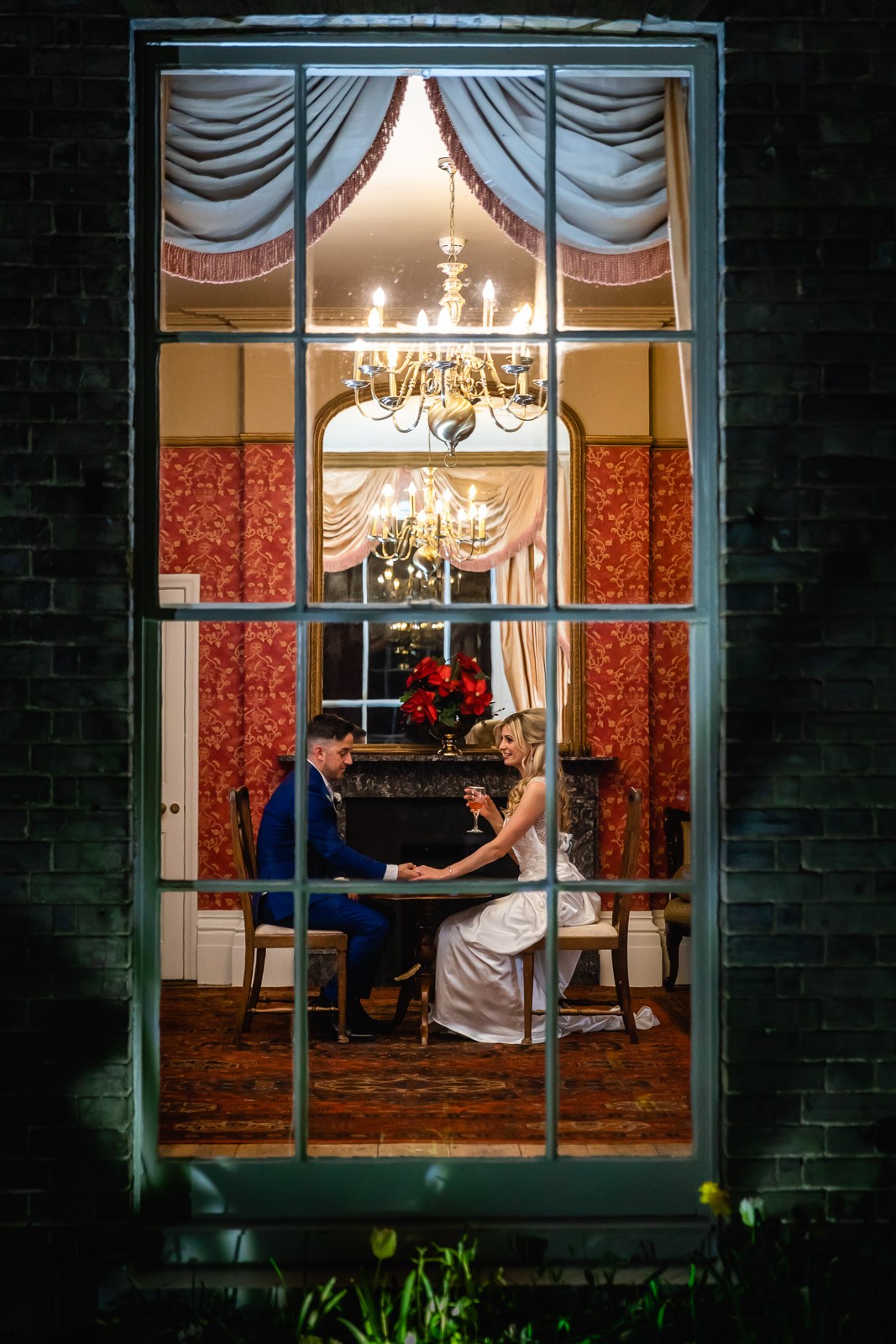 Through a window at night, the couple in the warmly lit chandeliered room at Fennes