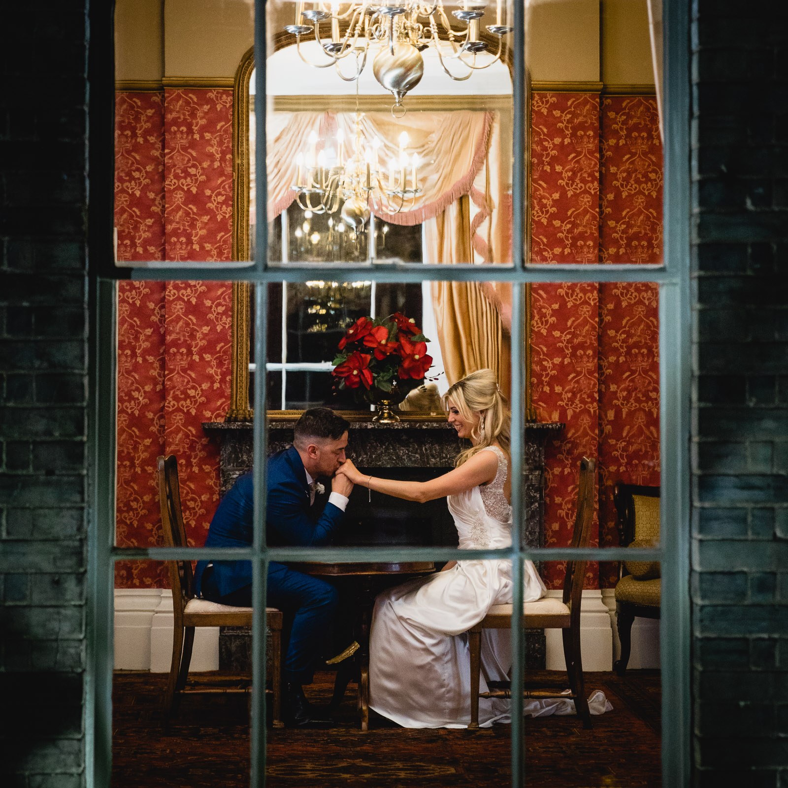 Through the window, the groom kissing the bride's hand at a candlelit table at Fennes