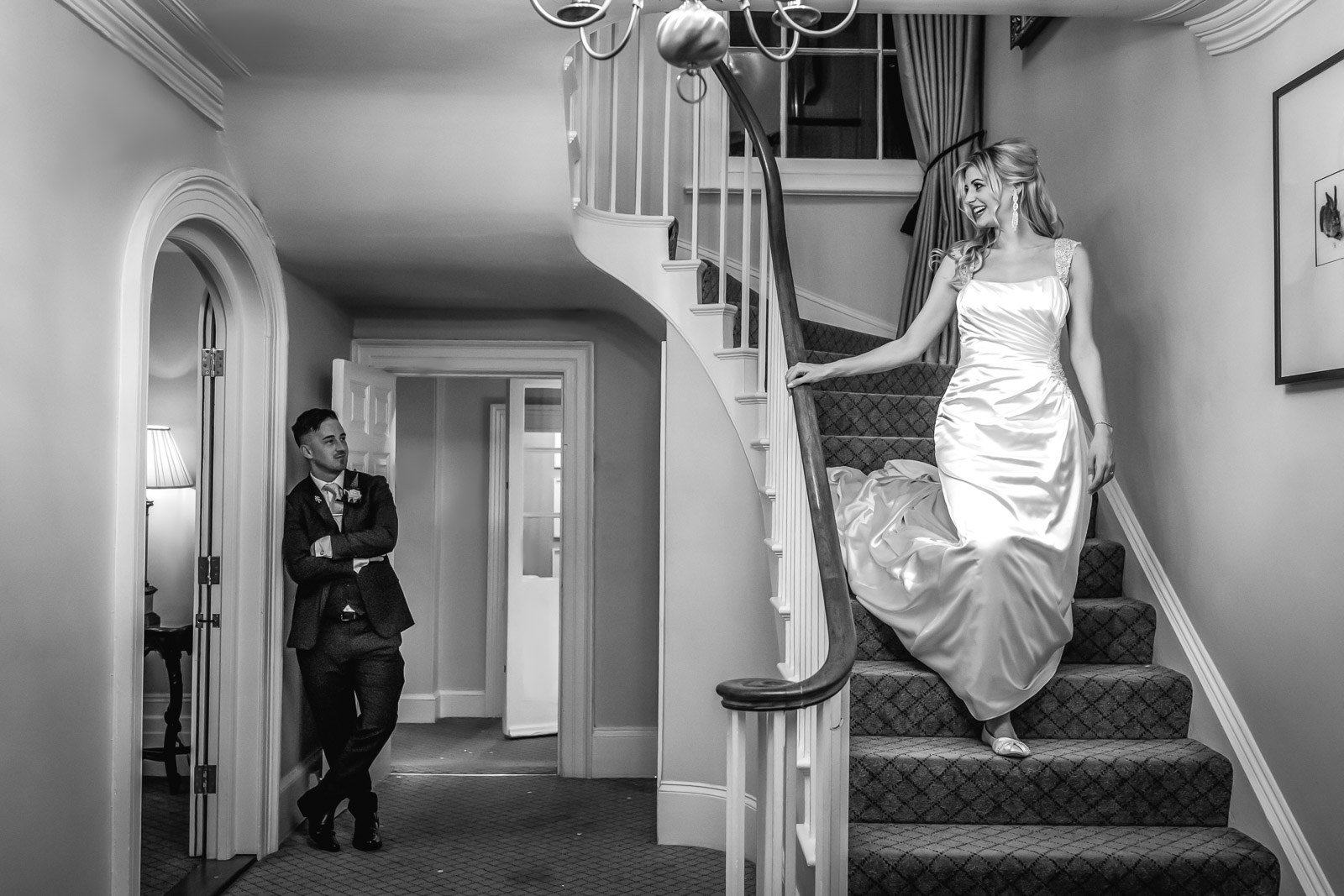 Black-and-white of the bride descending the sweeping staircase at Fennes, the groom below