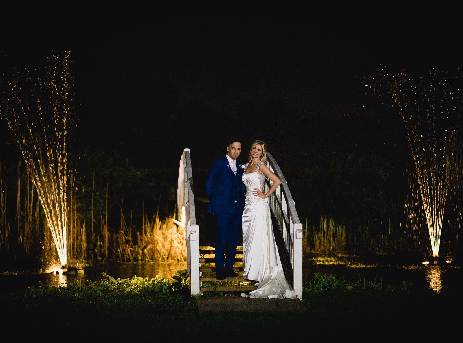 The couple on the footbridge at night, fountains lit behind them across the lake at Fennes