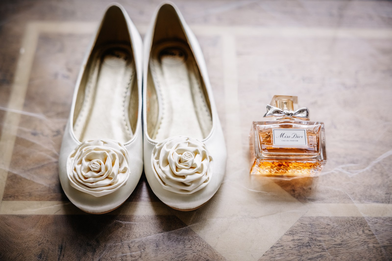 Ivory bridal shoes with satin rose detail beside a bottle of Miss Dior perfume