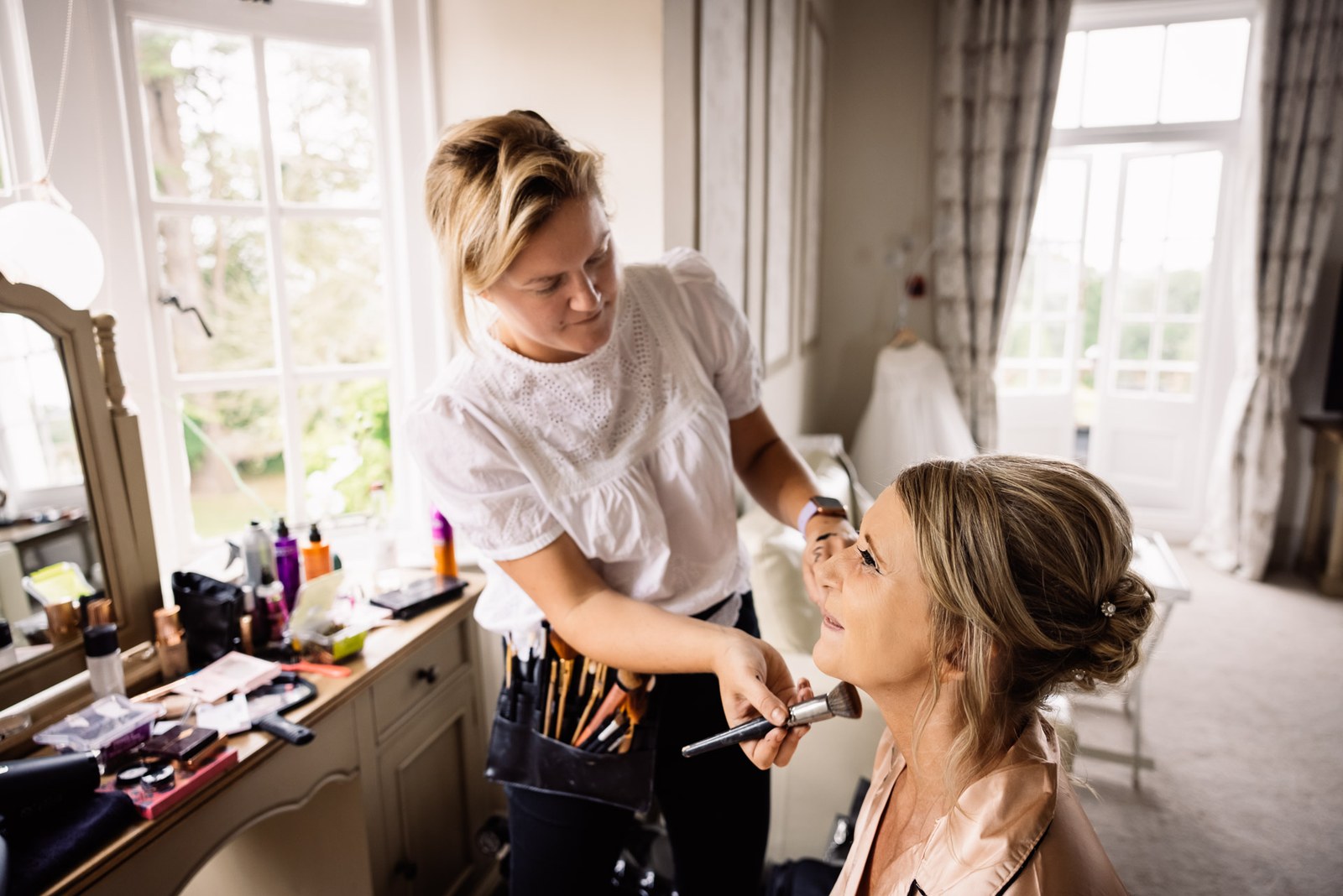 Makeup artist applying the bride's makeup by a sunlit bedroom window at Glenfall House