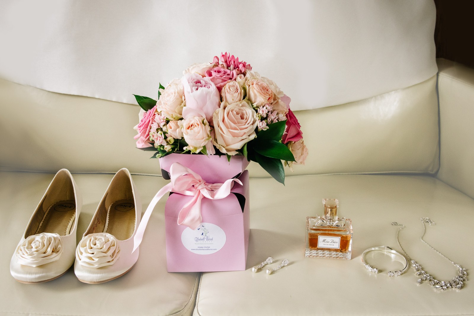 Bridal shoes, flowers, perfume and jewellery laid out on a cream sofa
