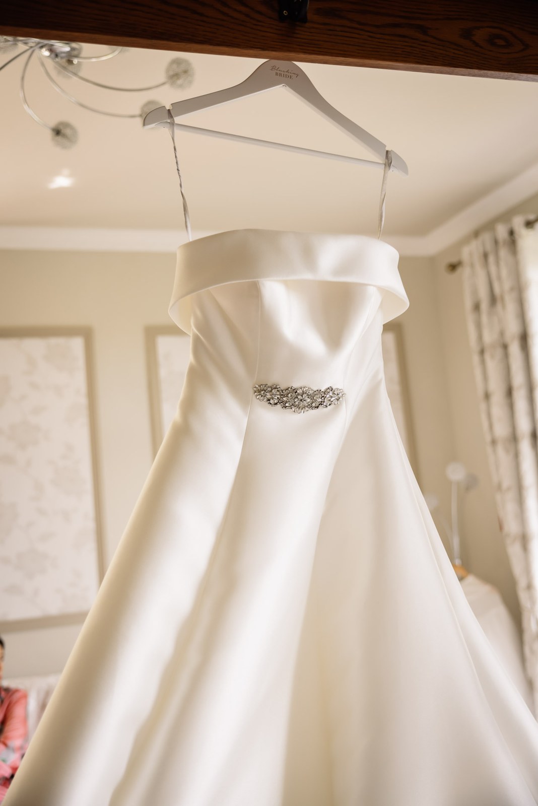 Strappy satin wedding dress hanging in a light-filled bedroom at Glenfall House
