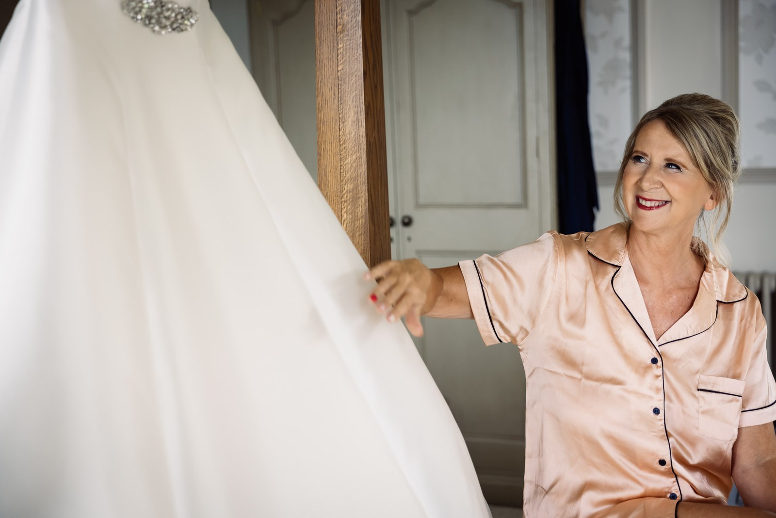 A smiling woman in pink pyjamas holding out the bride's hanging gown during the morning preparations