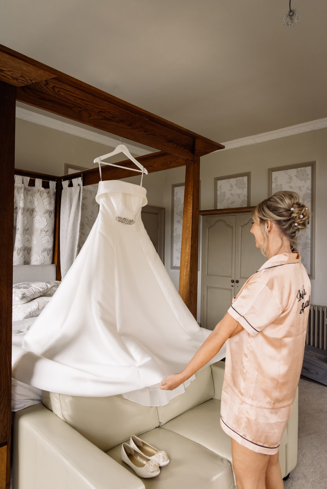 Bride smoothing the hanging wedding dress in a four-poster bedroom before the ceremony