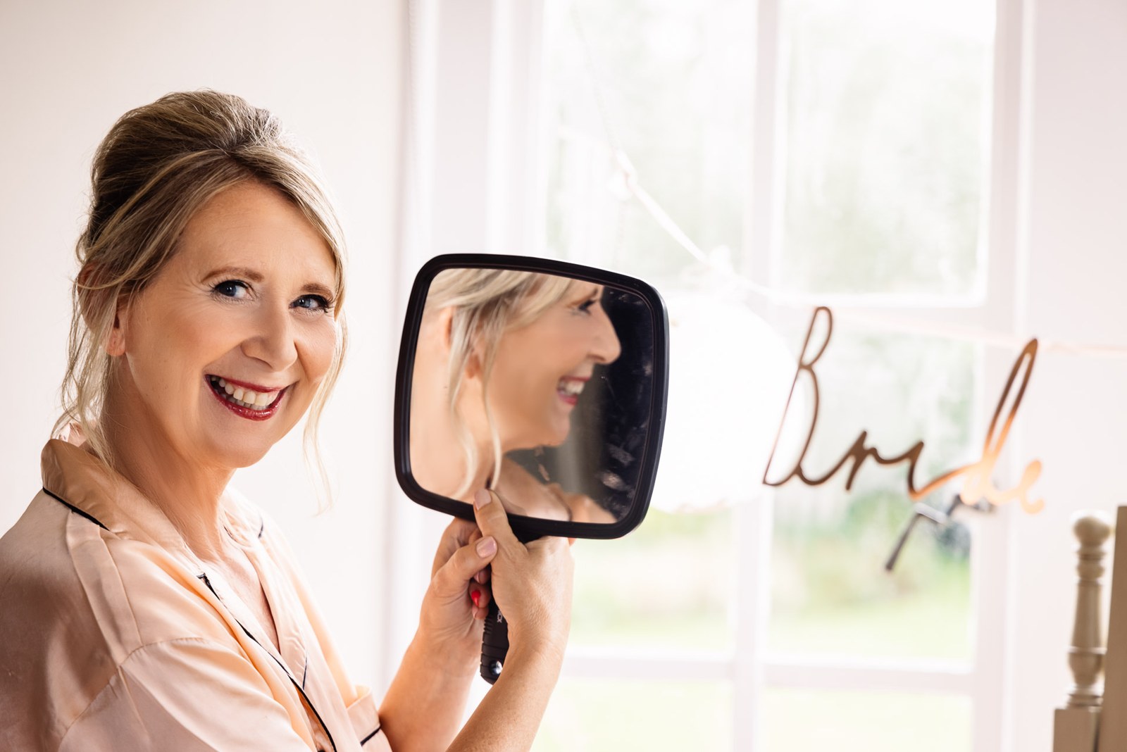 Bride laughing as she checks a hand mirror beside a gold 'Bride' sign