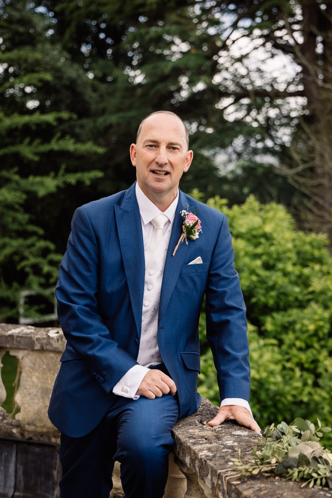 Groom in a blue three-piece suit relaxing on a stone garden wall at Glenfall House