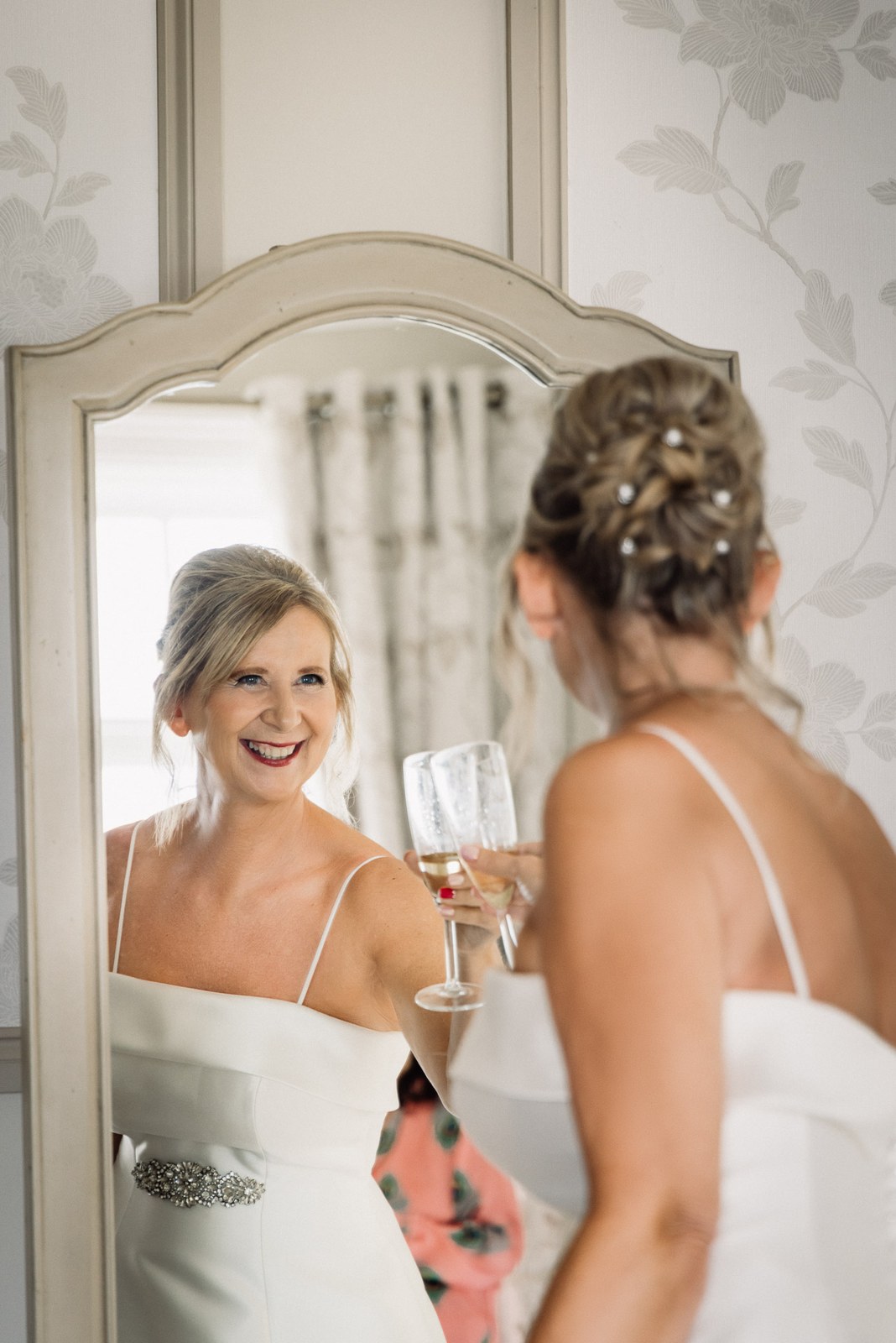 Bride holding a glass of champagne and admiring her gown in a bedroom mirror