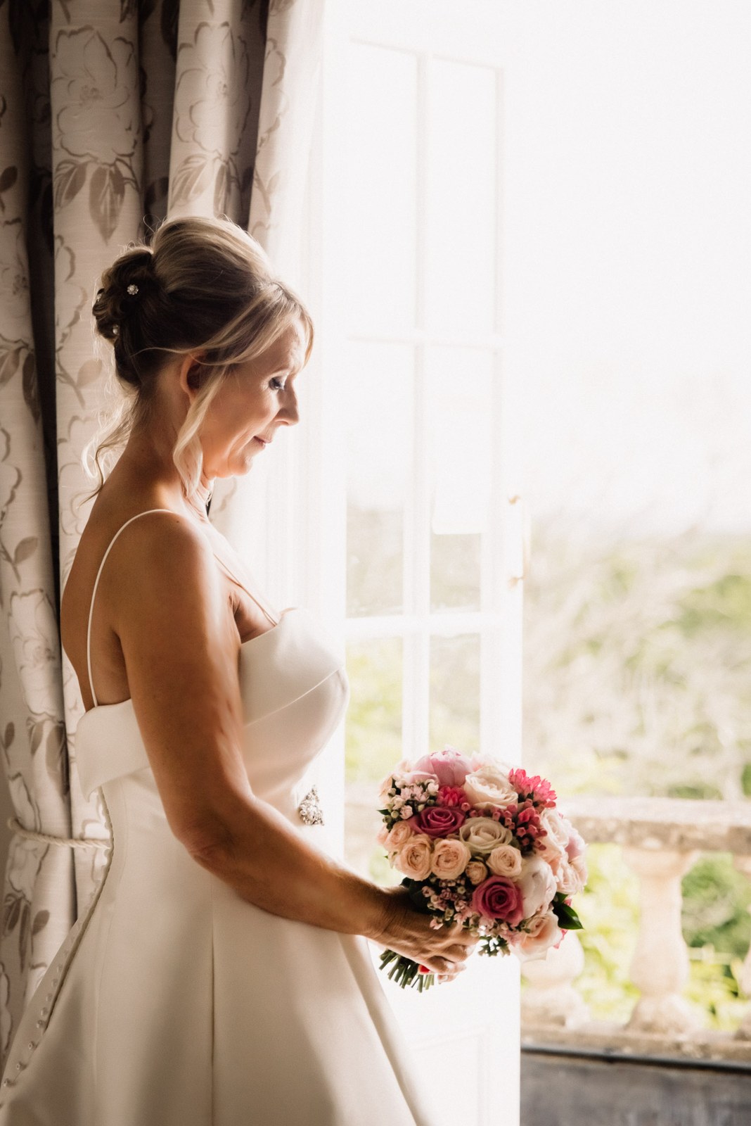 Bride standing at a tall window with her bouquet, soft daylight behind her