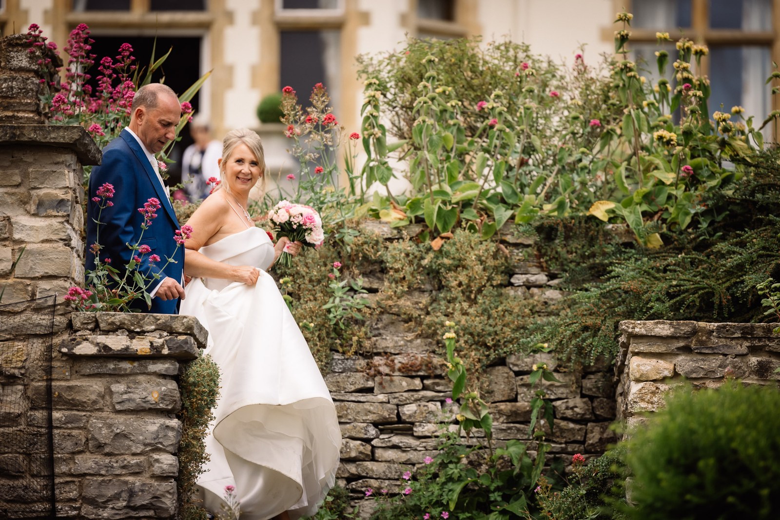 Bride and groom beside a dry-stone wall and cottage-garden borders at Glenfall House