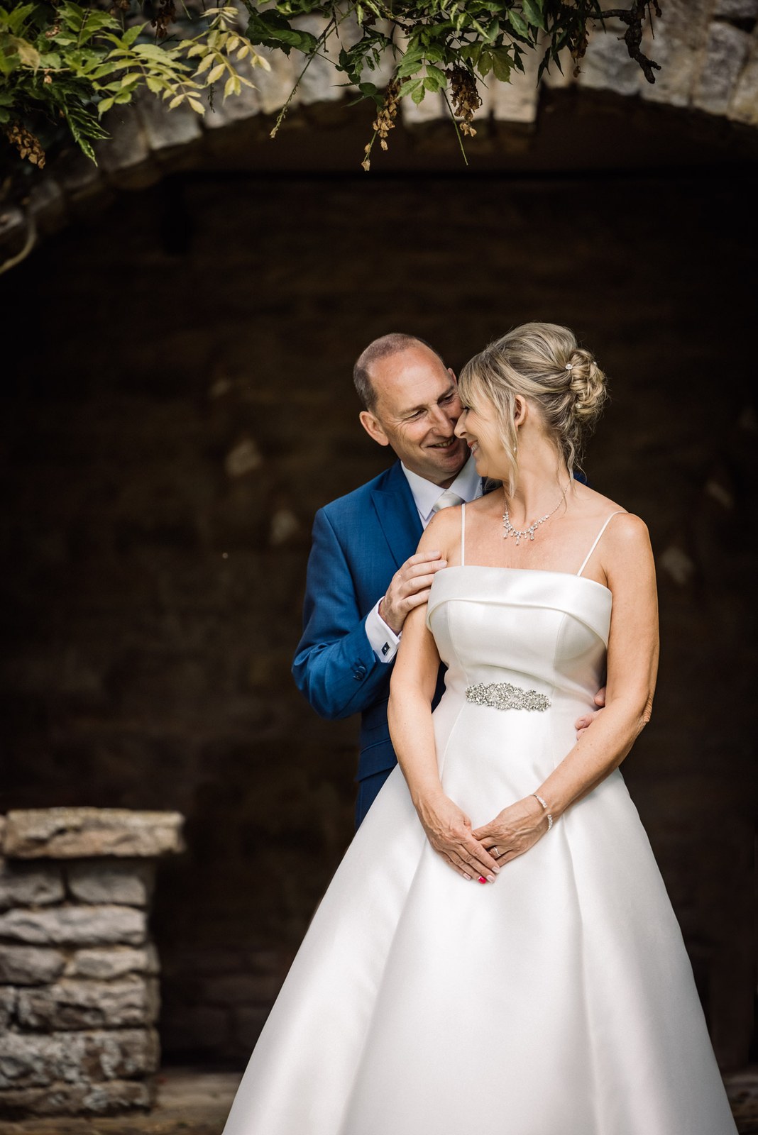 The groom leaning in cheek to cheek with the bride beneath the garden's stone archway