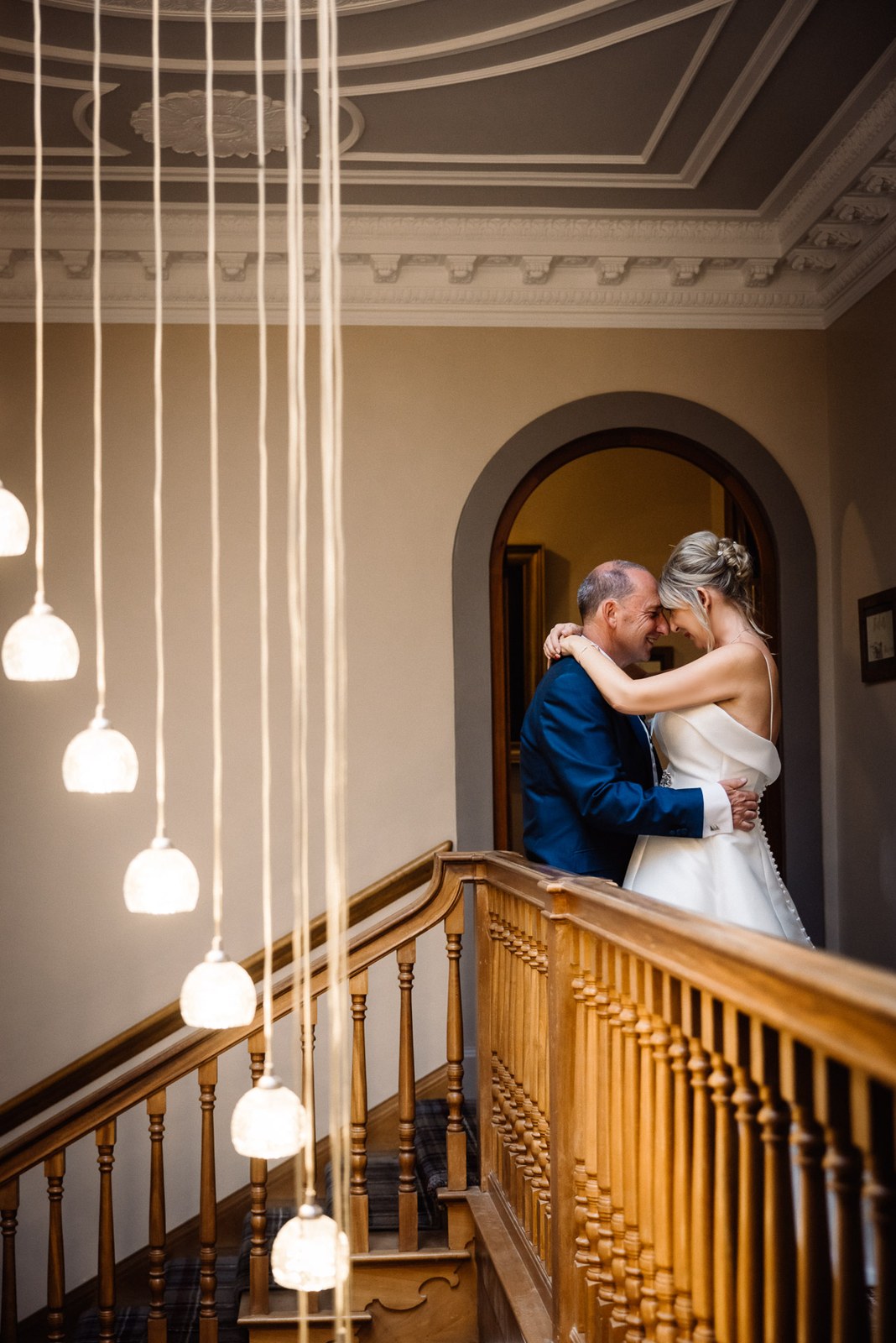 Bride and groom embracing on the staircase beneath Glenfall House's hanging bulb lights