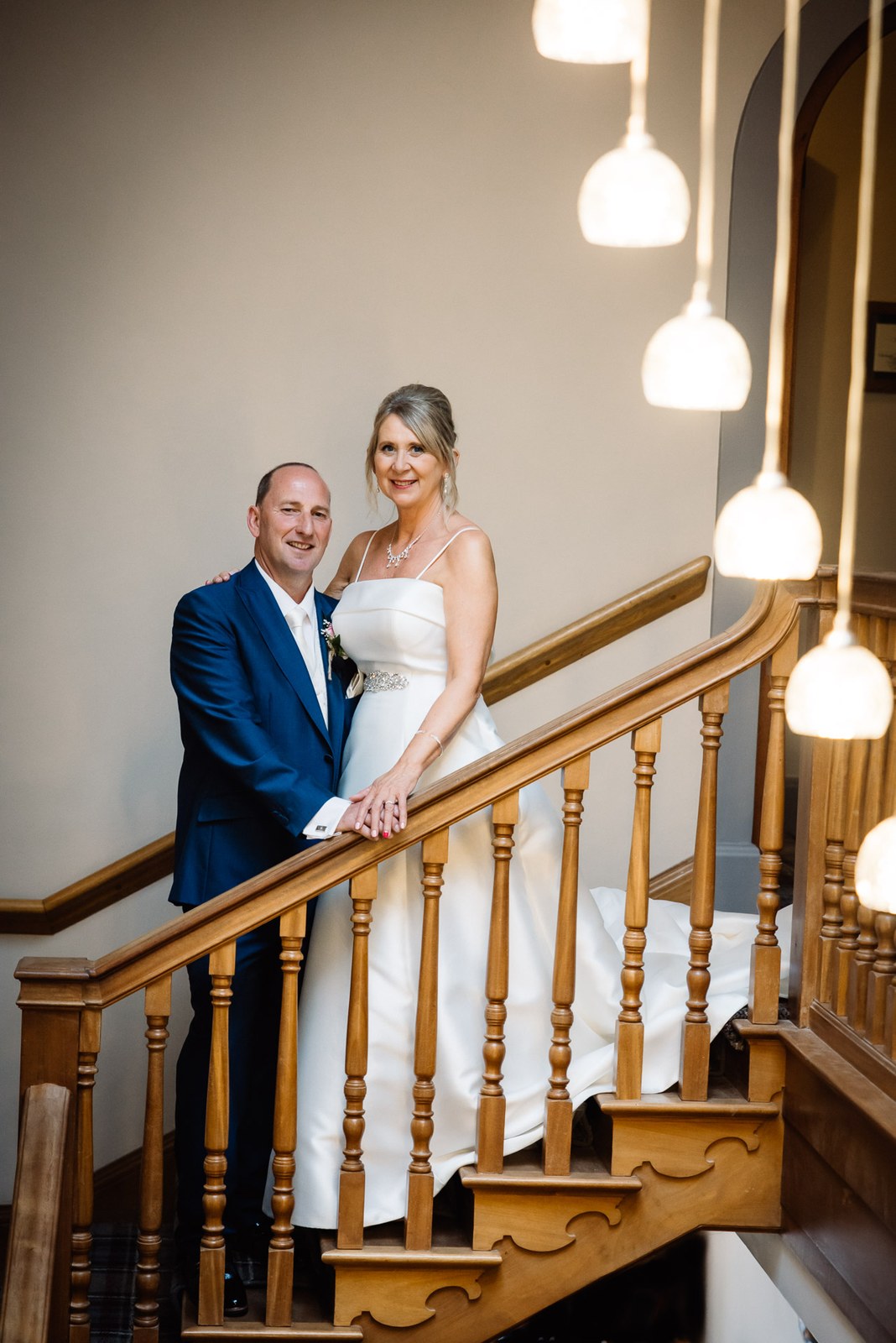 The newlyweds standing close on Glenfall House's grand wooden staircase