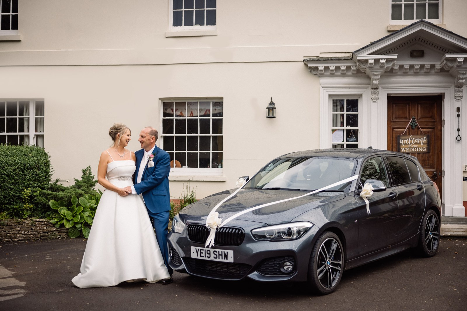 Bride and groom beside a ribboned wedding car at the entrance to Glenfall House