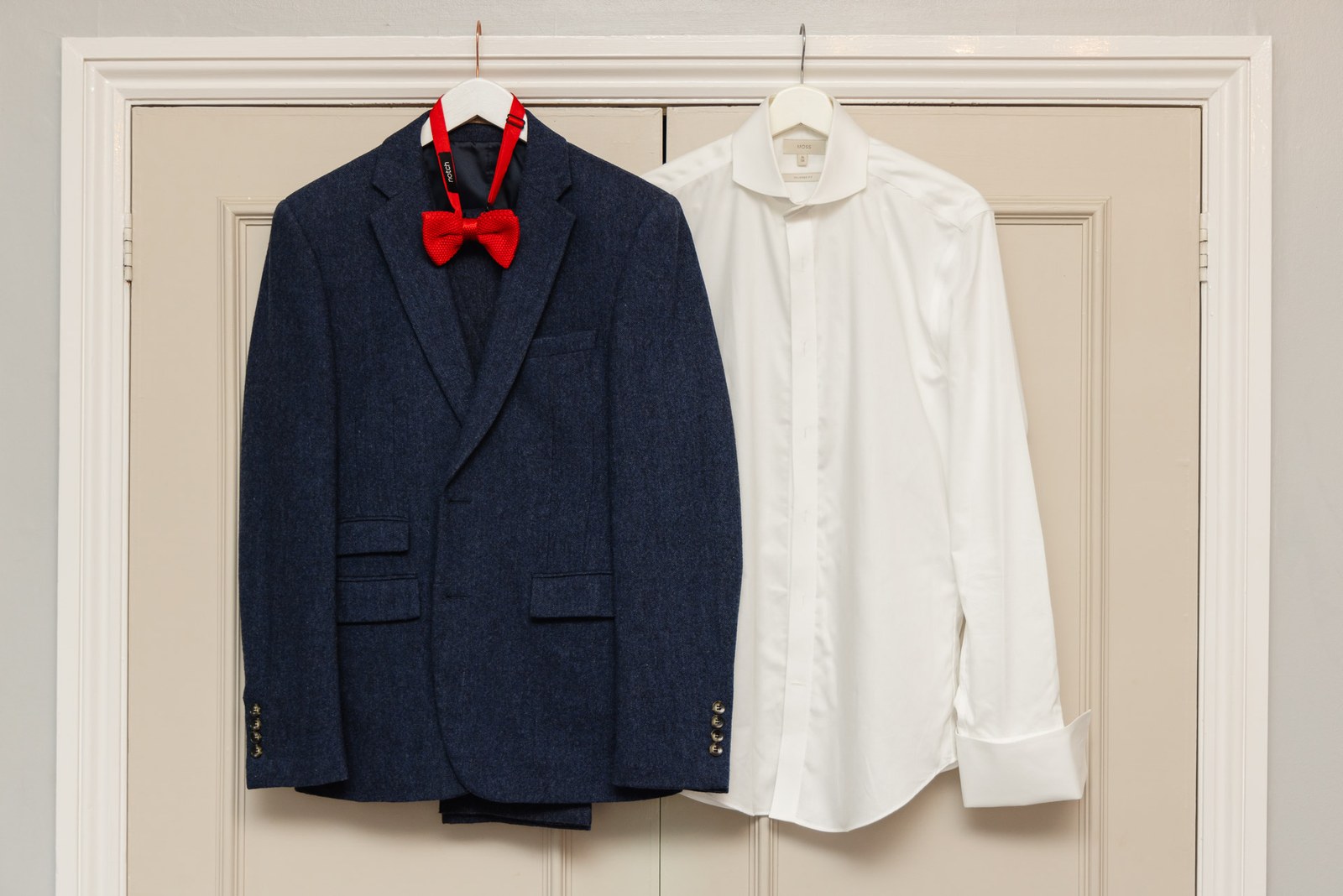 A navy suit jacket, white shirt and red bow tie hanging on a panelled door