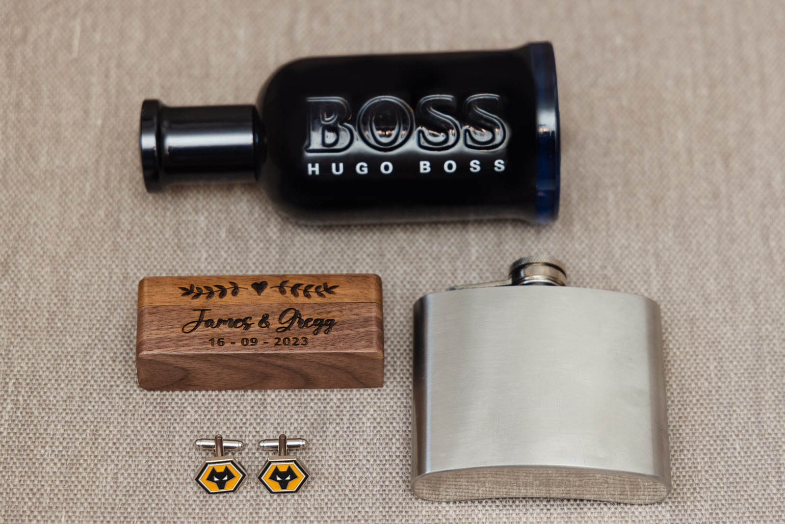 Flat lay of aftershave, an engraved wooden ring box, a hip flask and cufflinks