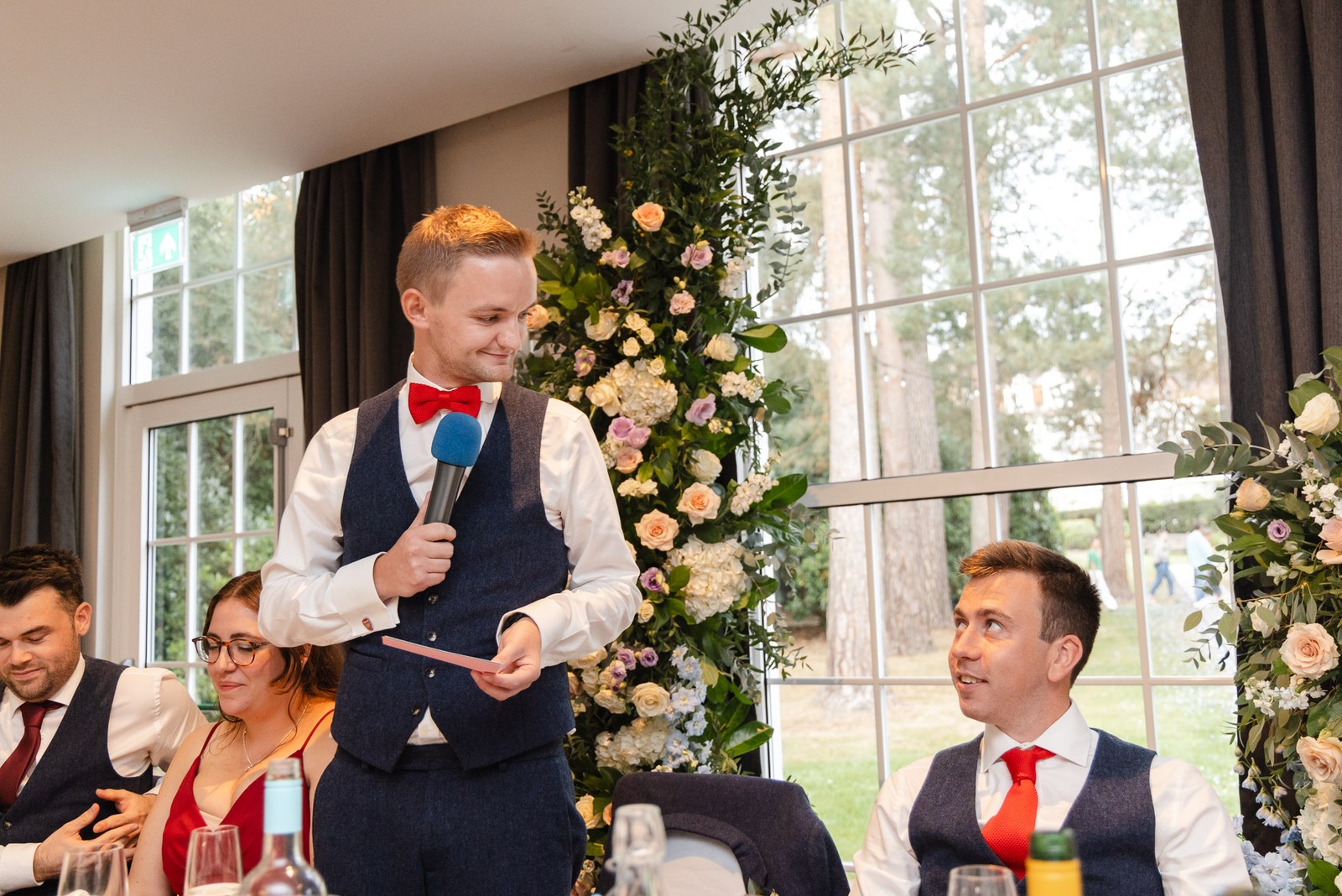 A groom delivering a speech with a blue microphone beside a floral arch