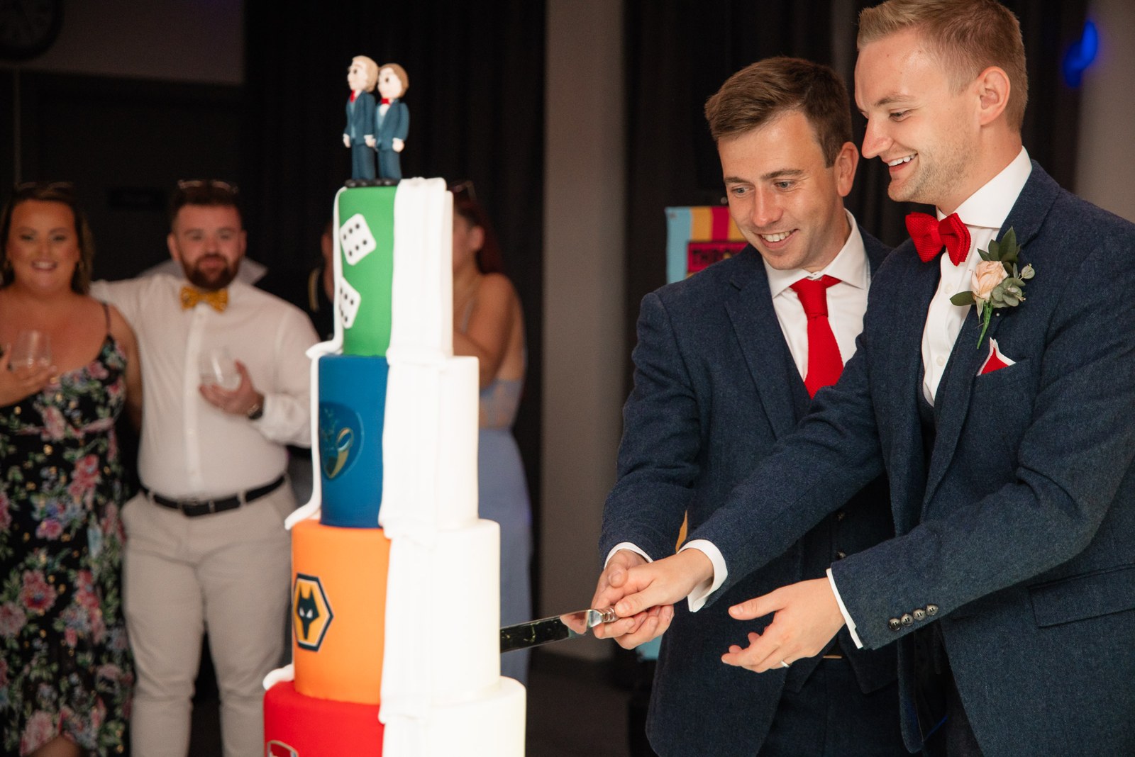 The grooms cutting a tall, colourful tiered wedding cake with a two-groom topper