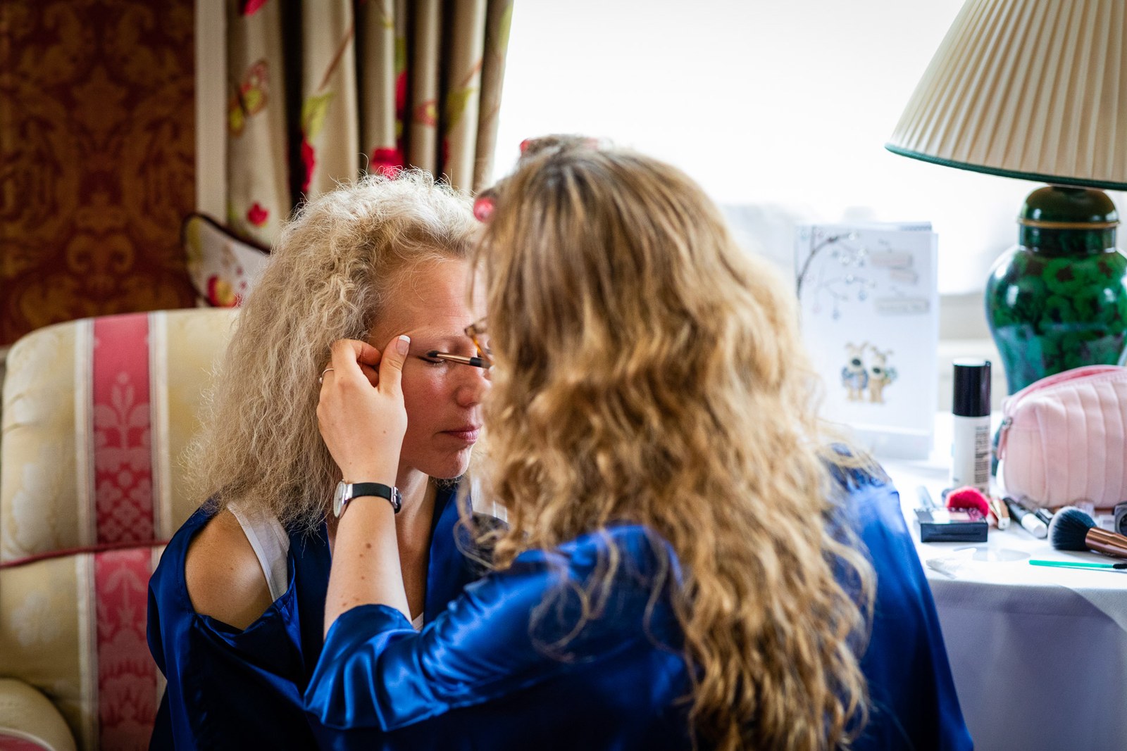 A bridesmaid in a blue robe helping the bride with her eye makeup