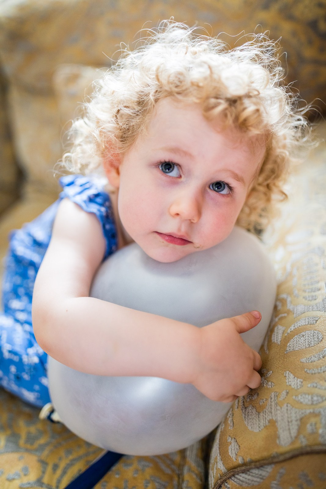 A flower girl in a blue patterned dress hugging a silver balloon in an armchair