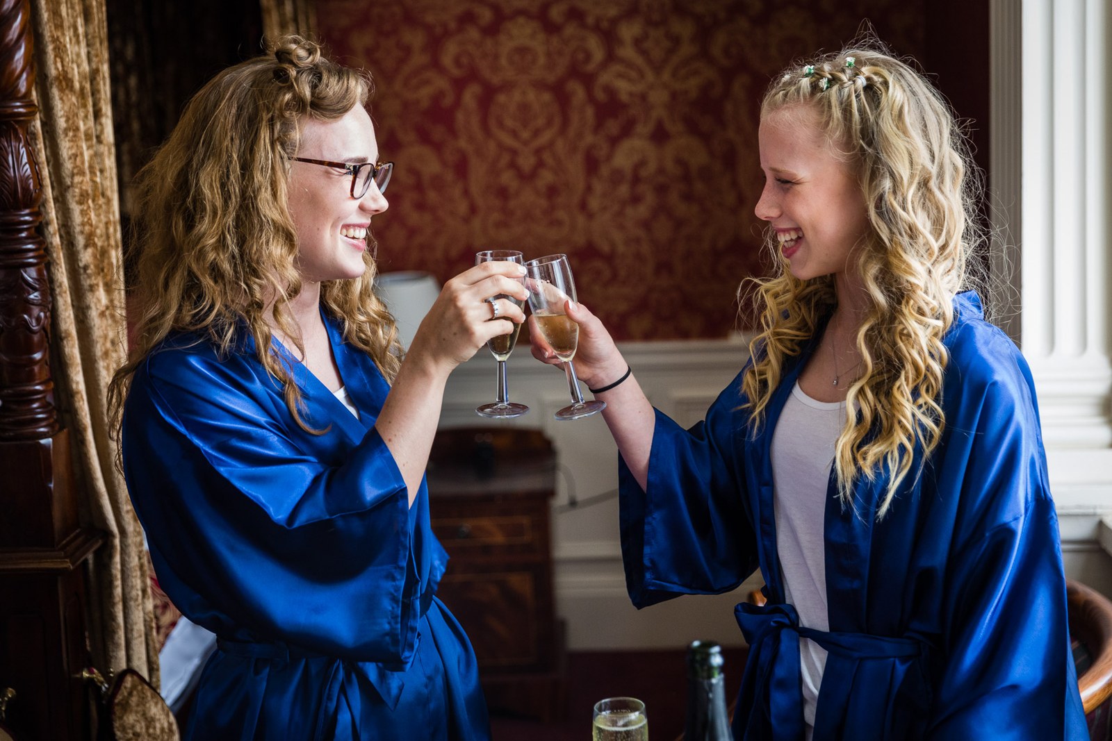 Two bridesmaids in blue satin robes toasting with champagne before the four-poster bed