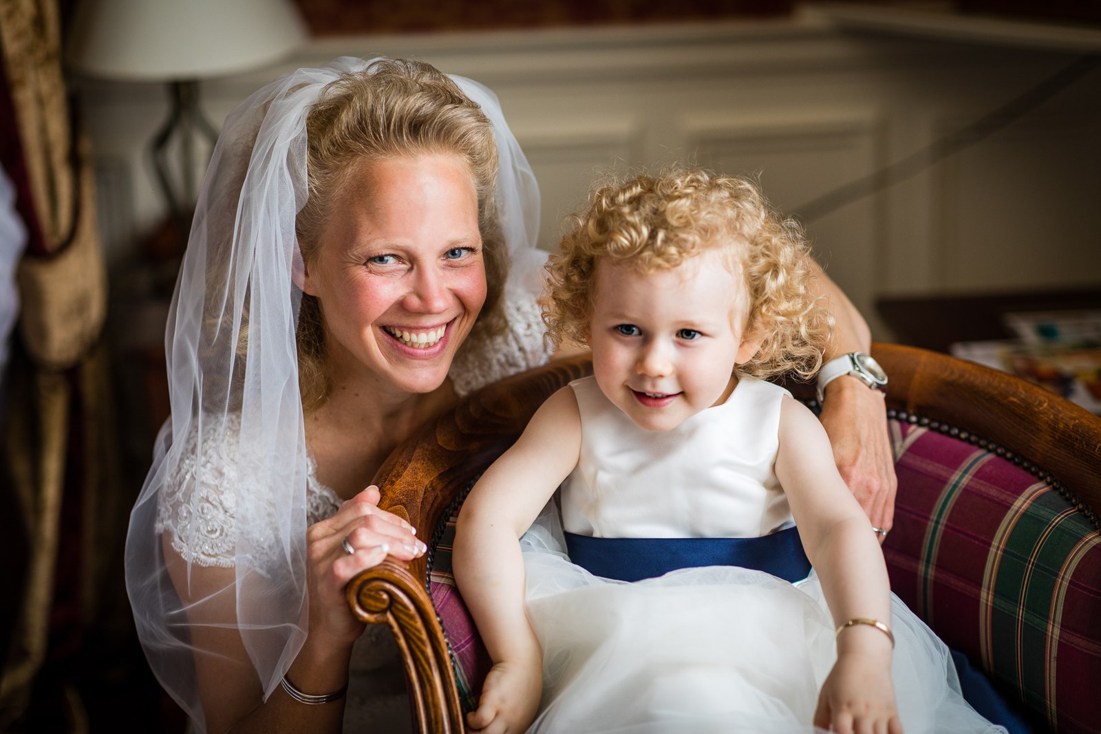 The bride cradling a flower girl on her lap, both smiling, in an armchair