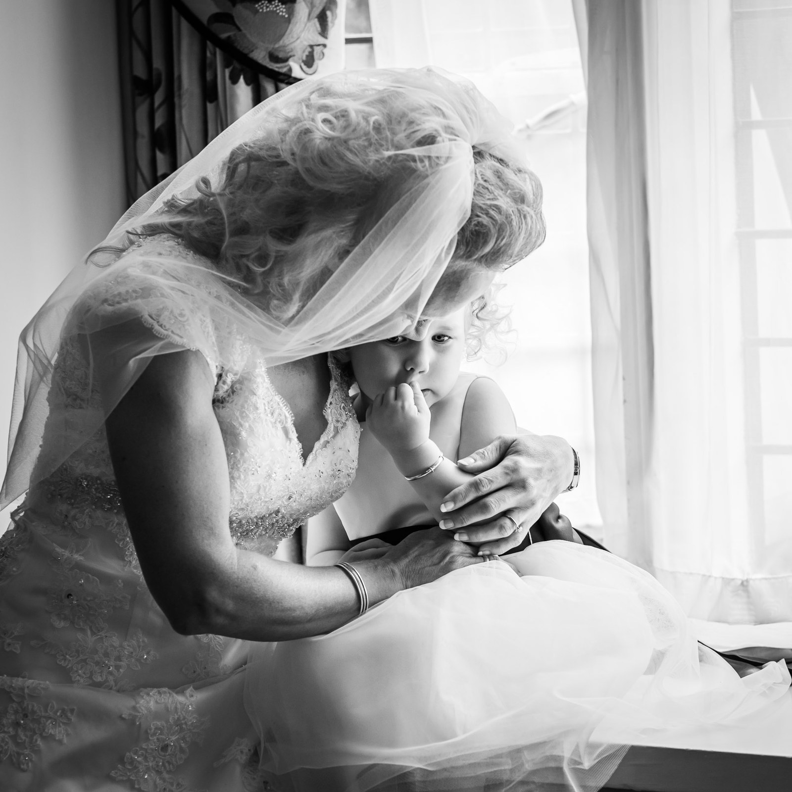 Black-and-white of the bride tenderly holding a flower girl's hand by a window