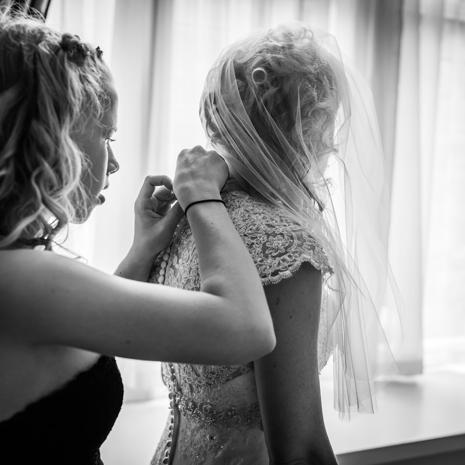 Black-and-white of a bridesmaid fastening a clasp at the back of the bride's lace gown