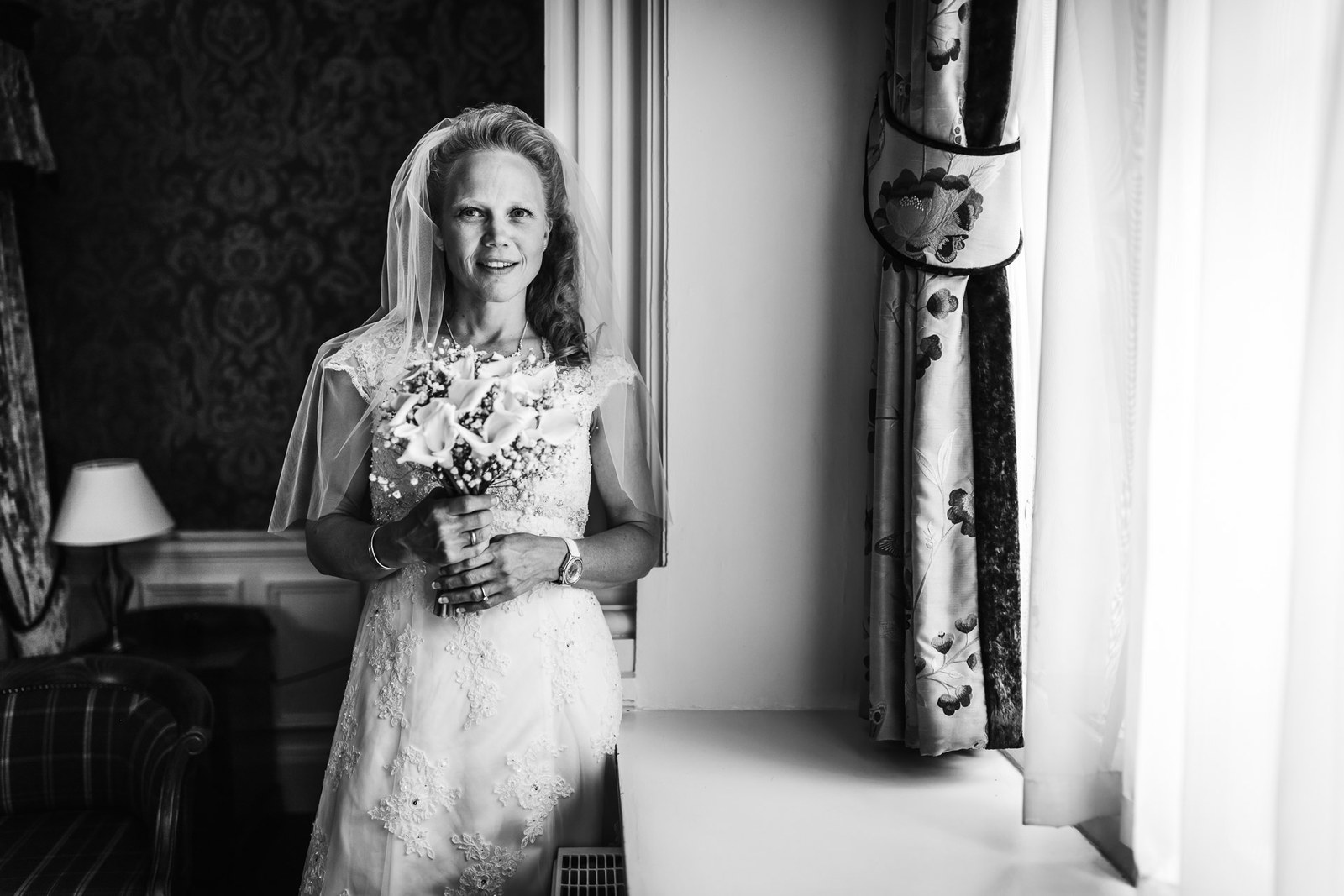 Black-and-white bridal portrait, the bride by a window in a damask-panelled room
