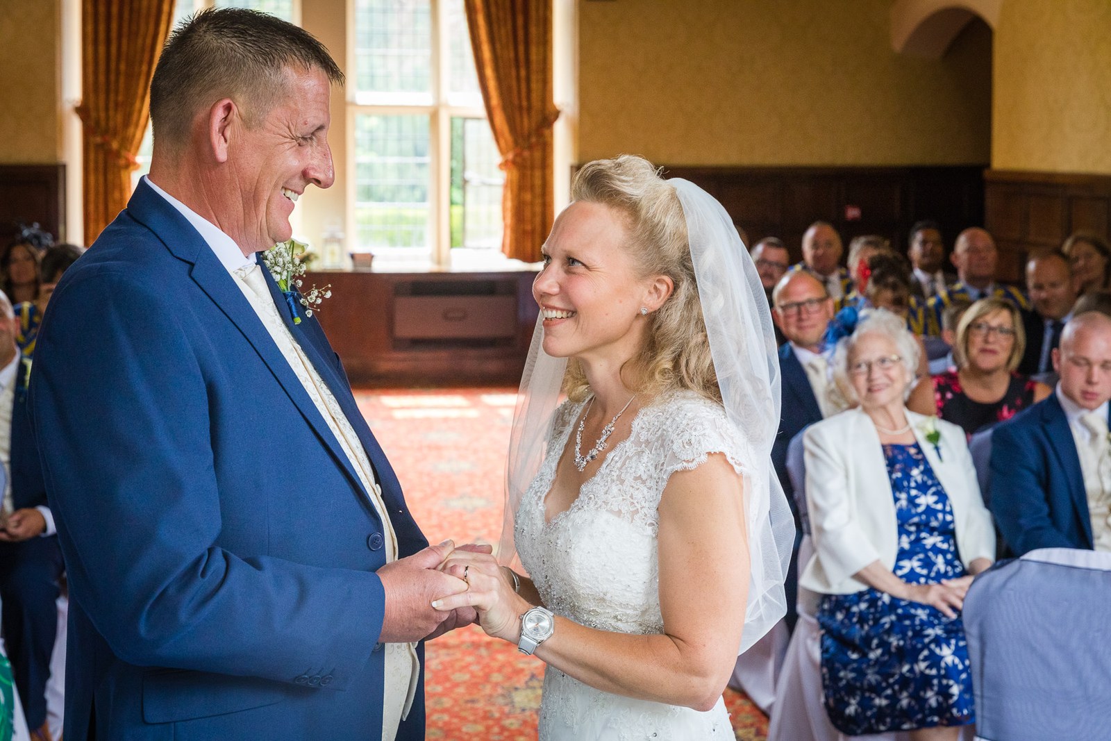 The couple holding hands during the ceremony in the panelled hall at Grim's Dyke Hotel, guests seated