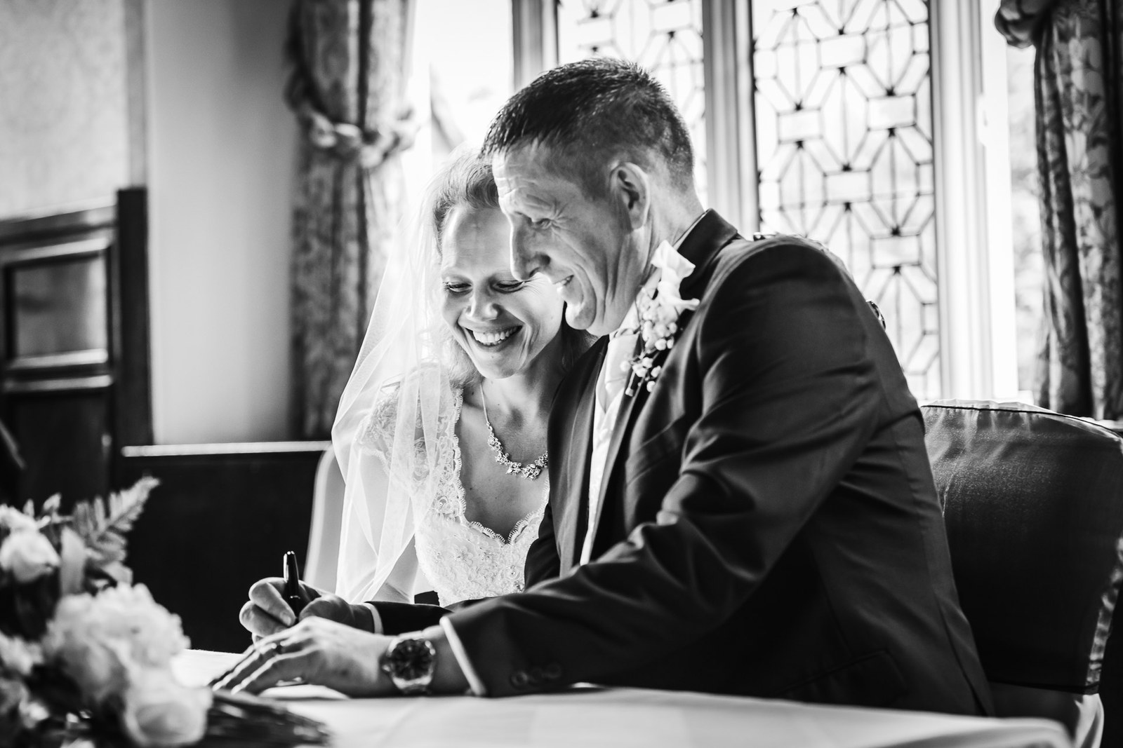 Black-and-white of the couple signing the register, the bride resting on the groom's shoulder