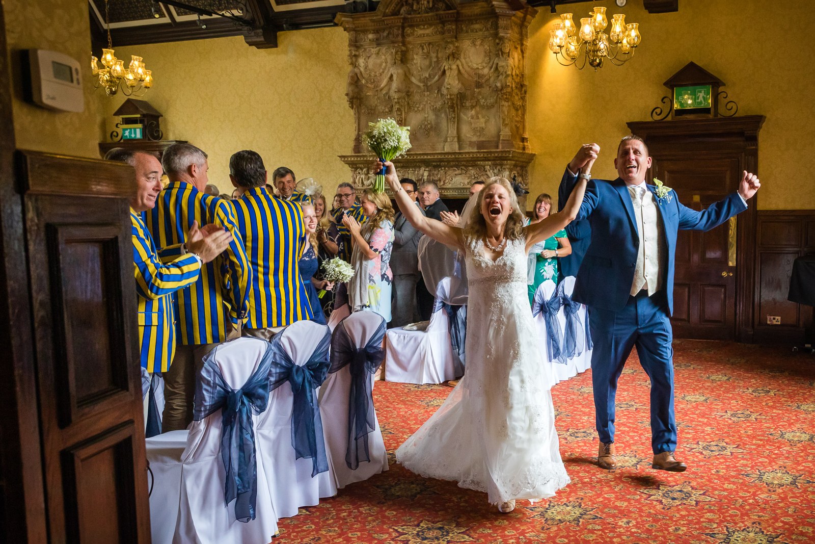 The couple celebrating with raised arms before the carved fireplace at Grim's Dyke Hotel, guests cheering