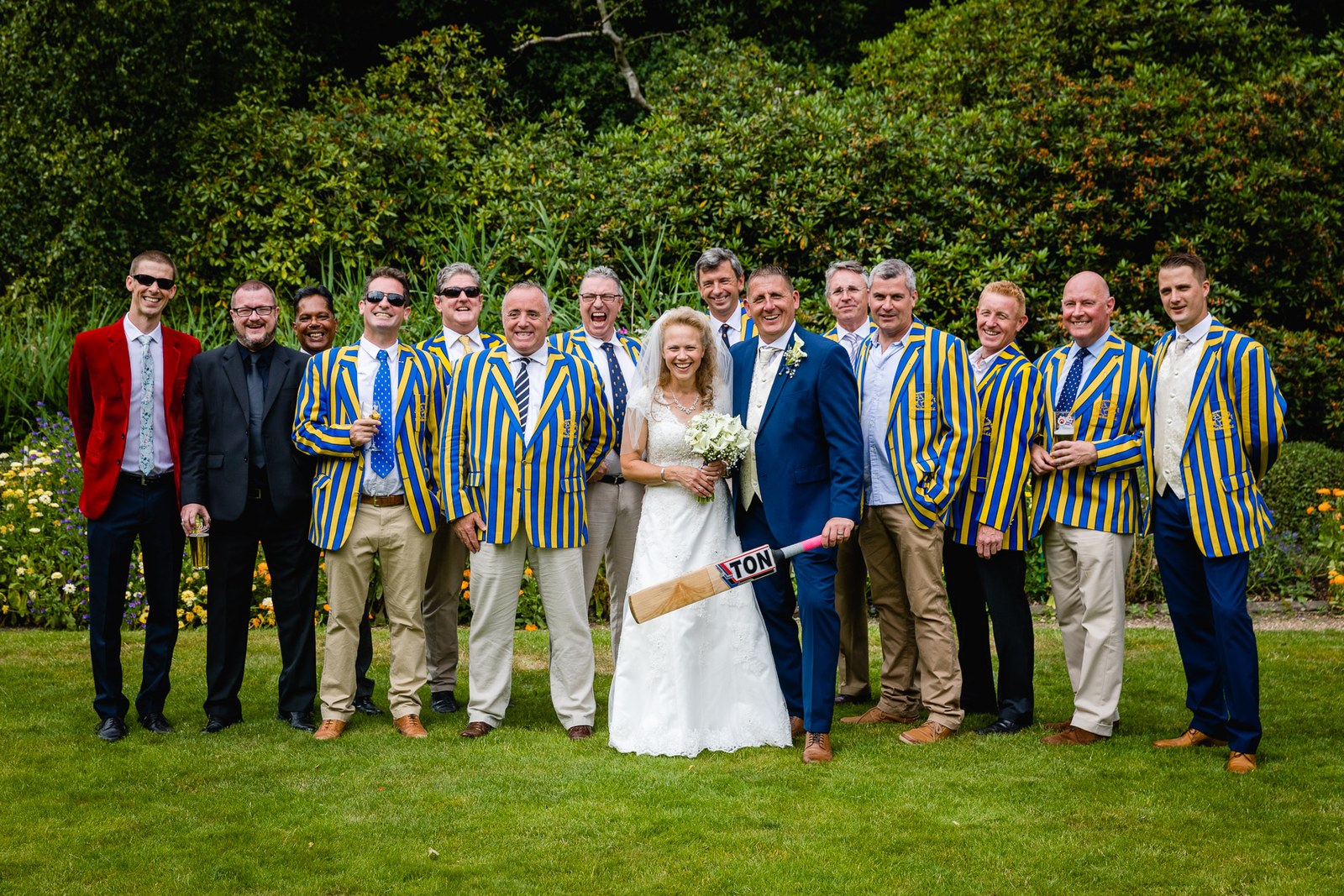 The bride with a group of men in striped cricket blazers holding a cricket bat on the lawn