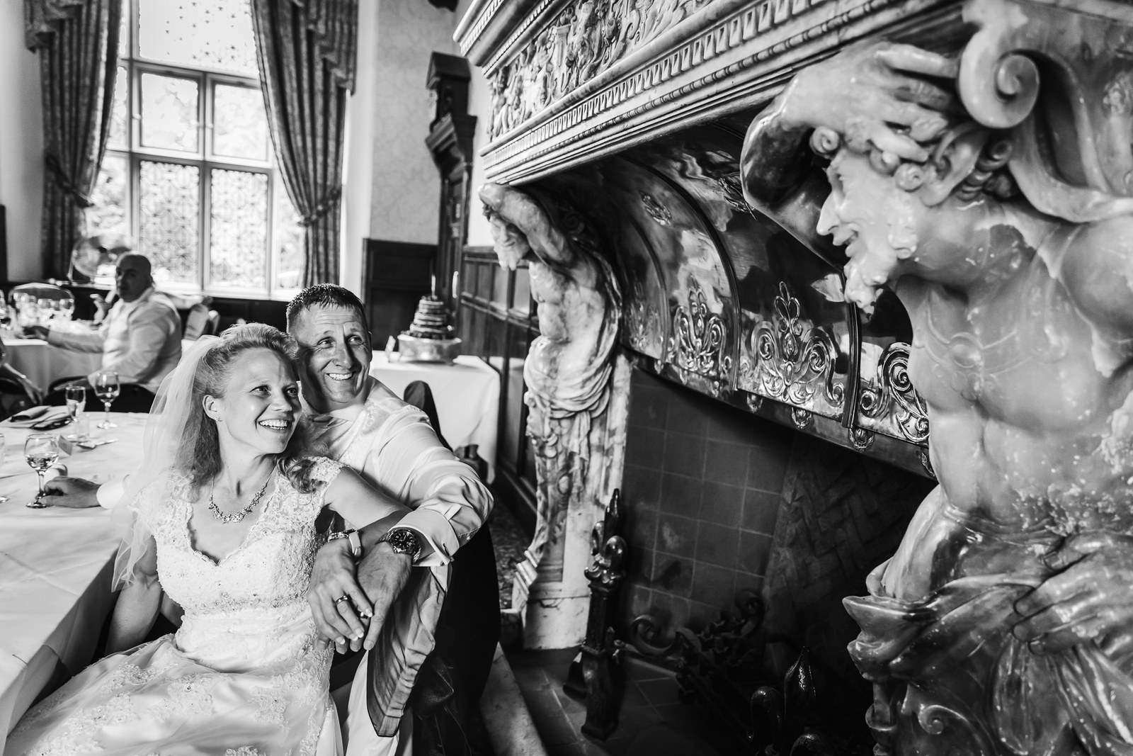 Black-and-white of the couple beside the elaborately carved figure fireplace at Grim's Dyke Hotel