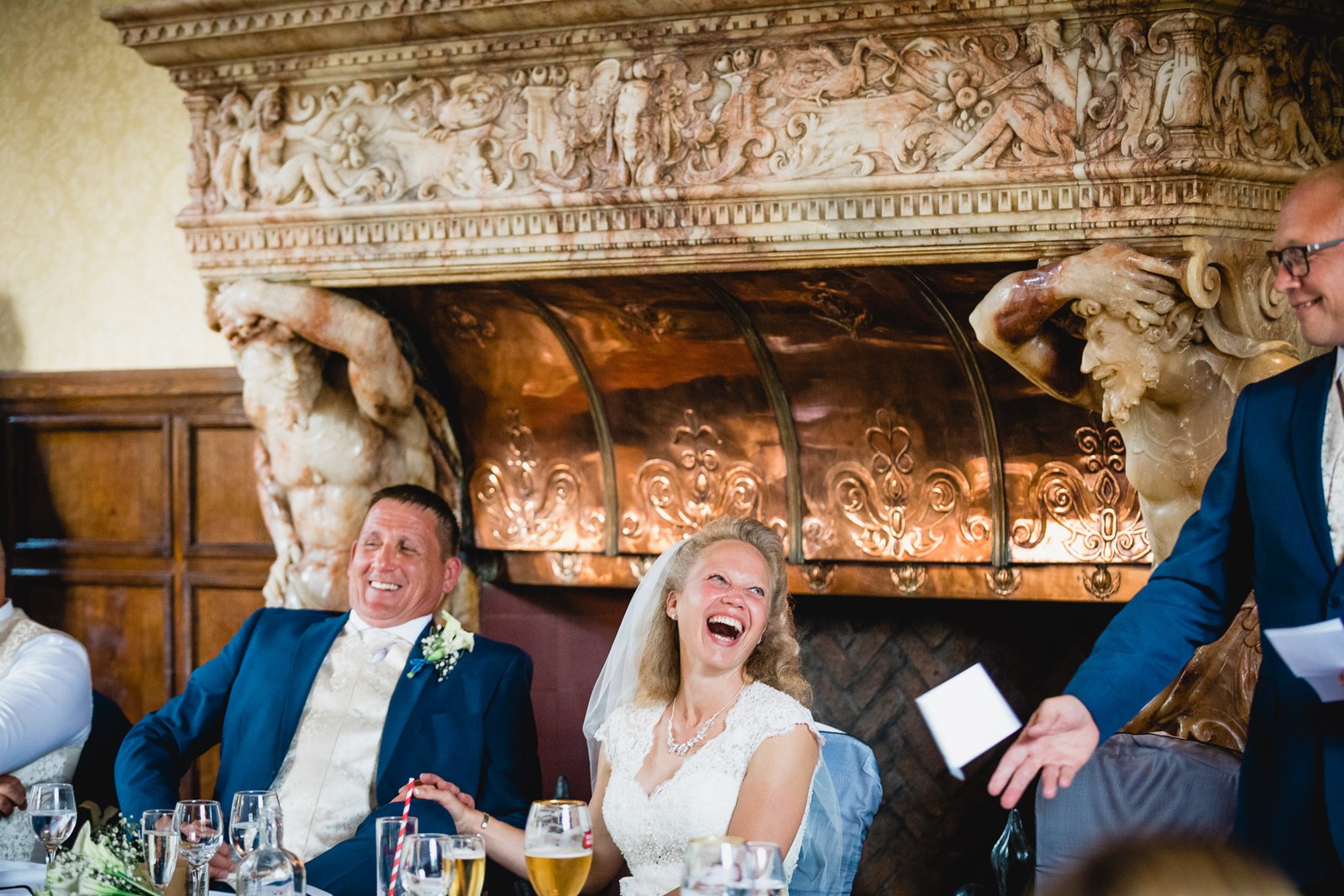 The couple laughing at the top table before the carved copper-hooded fireplace at Grim's Dyke Hotel
